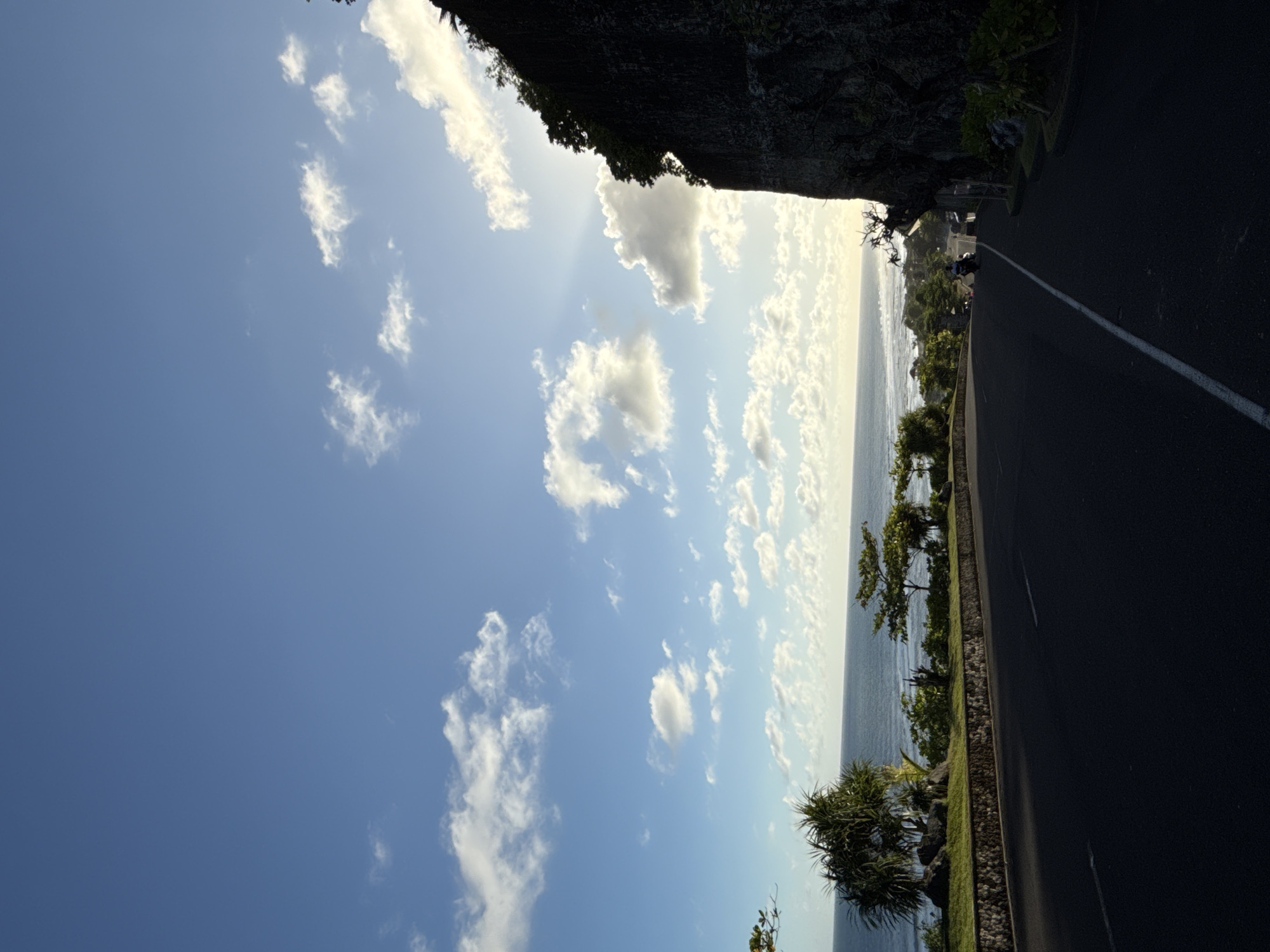 The image captures a coastal road scene in Ungasan, Indonesia. The view is angled with the sky occupying the majority of the left side of the frame. The sky is bright blue with scattered cumulus clouds, some illuminated by the sun behind a cliff face to the top right. The ocean is visible along the right edge of the image, separated from the road by a low wall and sparse vegetation including small trees and bushes. A dark asphalt road runs along the right side, with a faded white line marking the center. In the distance, figures on motorcycles are faintly visible on the road. A cliff or raised terrain with dense foliage borders the top of the image.