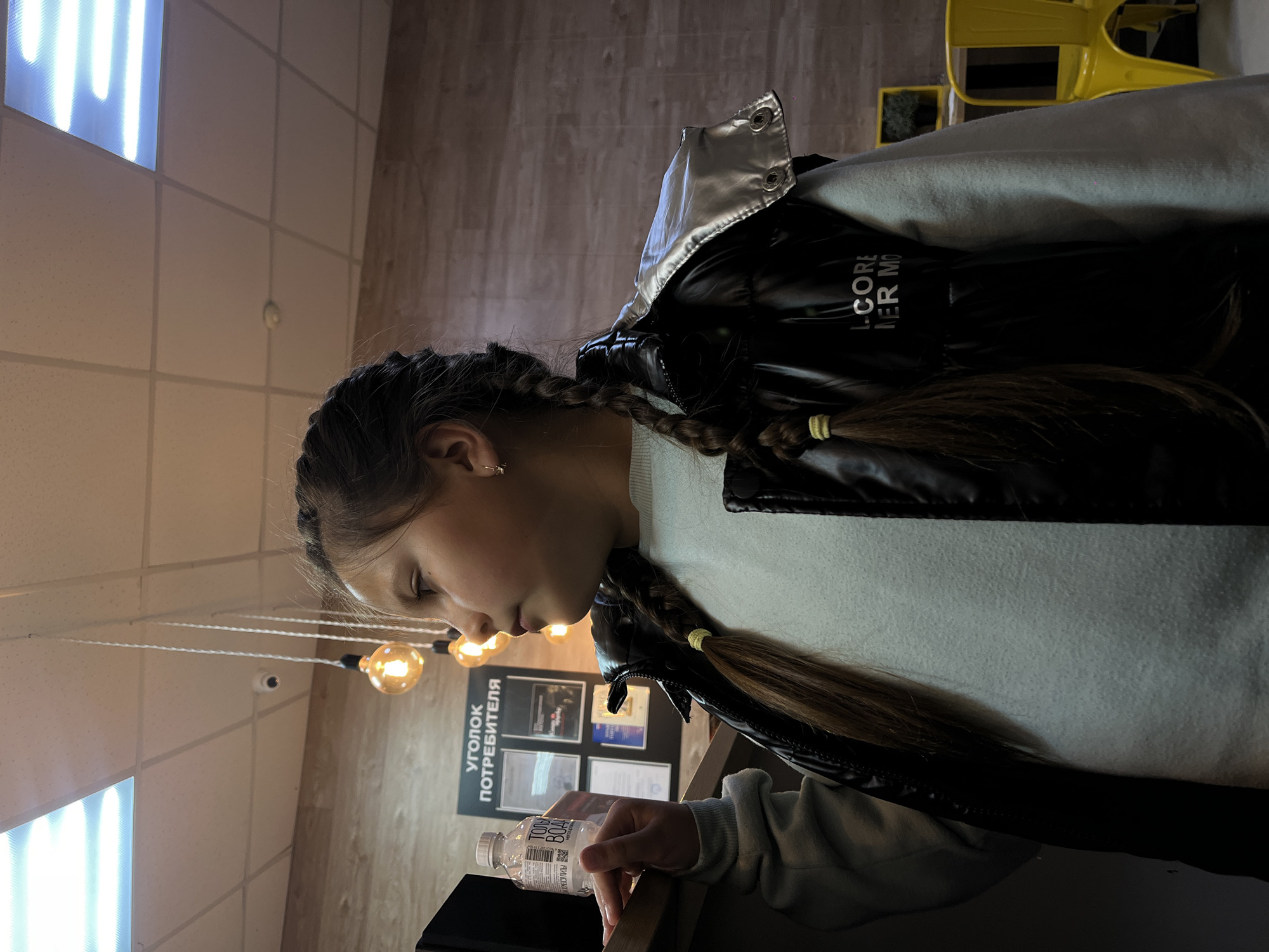 A young girl, likely a pre-teen or early teen, is the central subject of this indoor, sideways-oriented photograph taken in Novopokrovskaya, Russia. She is shown in profile, looking downwards with a neutral expression, holding a clear plastic water bottle in her right hand. Her hair is styled in two braids, secured with yellow elastics, and she is wearing a light-colored long-sleeved top beneath a black puffer vest with a shiny silver collar. The vest has partially visible text that reads "L. CORE" and "MER MO".
The setting appears to be a commercial or public indoor space, possibly a cafe or shop. In the background, a sign is prominently displayed on the wall, reading "УГОЛОК ПОТРЕБИТЕЛЯ" (Consumer's Corner), with several documents or certificates arranged beneath it. Various light sources illuminate the scene, including bright overhead fluorescent panels and a cluster of hanging Edison-style bulbs. A small security camera is visible on the ceiling. In the far background, part of a yellow chair can be seen, along with what appears to be a plant in a dark pot. The walls feature a tiled pattern, and a wooden texture is visible, possibly on the floor or another wall. The water bottle in the girl's hand has a label that clearly shows "ТОНУС ВОДА" (TONUS VODA) and "НЕГАЗИРОВАННАЯ" (non-carbonated).