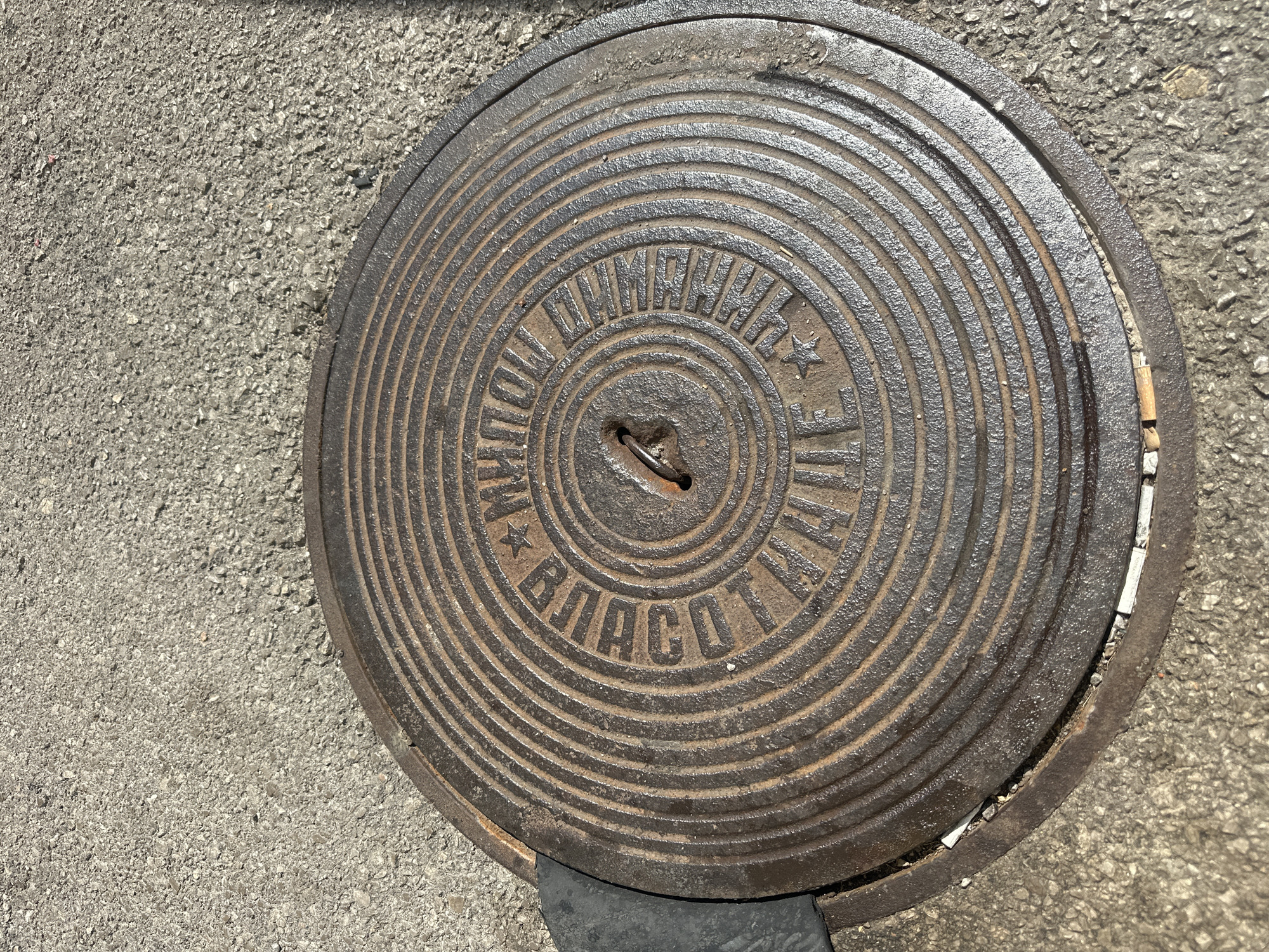 A weathered, circular cast-iron manhole cover, partially ajar, rests on a dark grey asphalt surface. The manhole cover is adorned with concentric ridges and raised lettering. The lettering appears to be in Cyrillic script, with prominent words like "ВОДА" (water) and "КАНАЛИЗАЦИЯ" (sewerage) visible. A small, rusted metal loop is present in the center of the cover, likely for lifting. A dark rubber or plastic object, possibly a gasket or part of a lifting tool, is partially visible to the left of the manhole cover, wedged between it and its frame. Scattered debris, including small stones and what appear to be cigarette butts, are present on the asphalt around the manhole cover. The scene is illuminated by direct sunlight, casting subtle shadows and highlighting the texture of the asphalt and the metal cover. The location is the city of Budva, Montenegro.