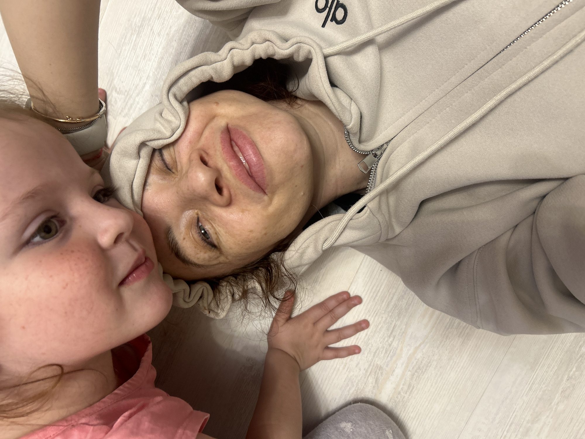 A woman and a young girl are lying on a light-colored wooden floor. The woman is wearing a beige hoodie with the logo "Alo" visible. The girl is wearing a pink shirt and has reddish-blonde hair and freckles. The woman's left hand is near the girl's face. The setting appears to be indoors, possibly in a private residence. The image was taken in Chaikovsky, Russia.