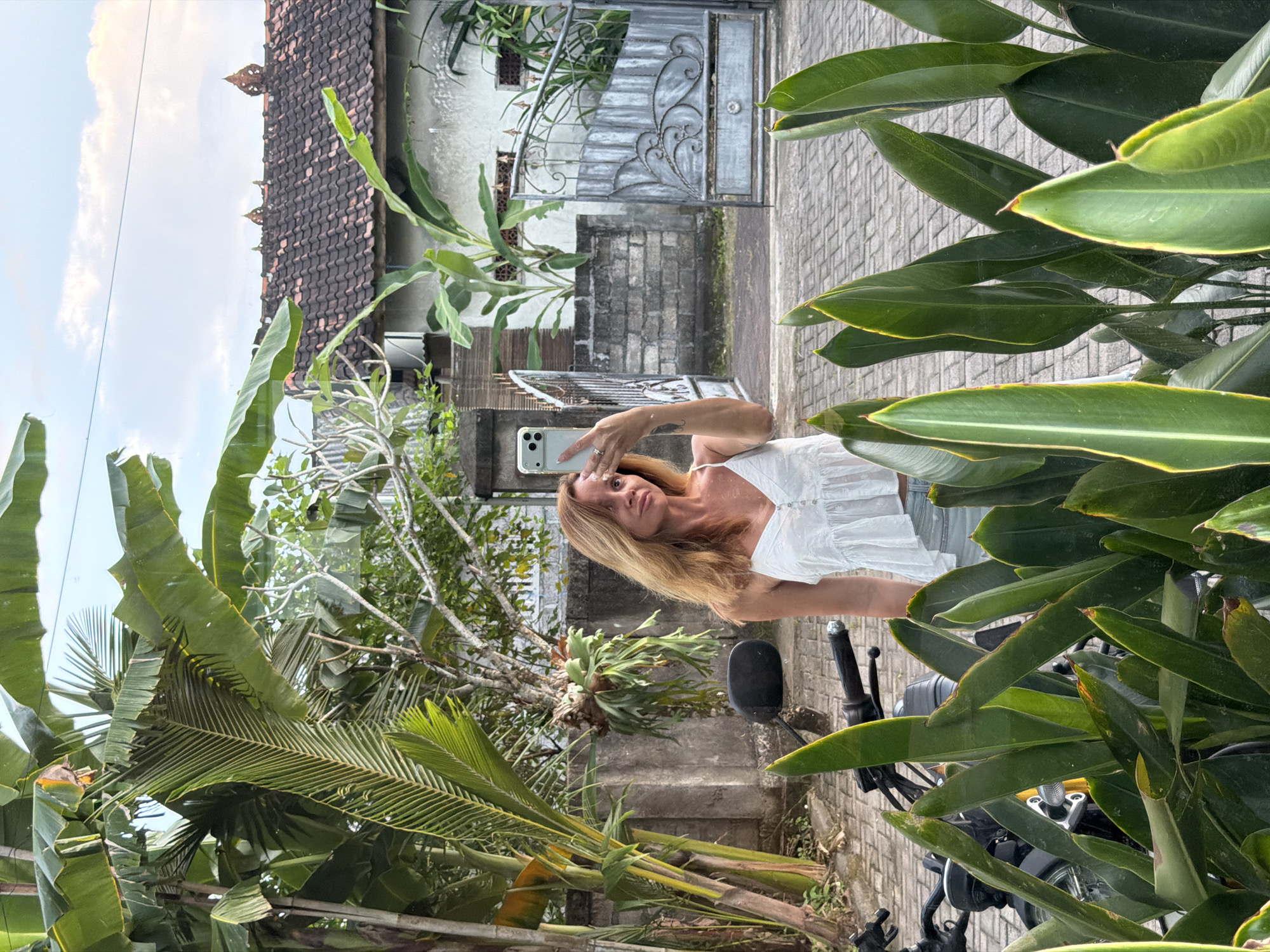 A woman stands outdoors, holding a smartphone up, likely taking a selfie. She is framed by lush tropical foliage, including banana trees and other large-leafed plants. Behind her is a residence with a tile roof and a gray brick wall topped with a decorative iron gate. The setting appears to be a street paved with interlocking bricks, with a motorcycle partially visible in the foreground on the right. Given the proximity to tropical plants and similar residential architecture, the location is likely Kuta Selatan, Indonesia.