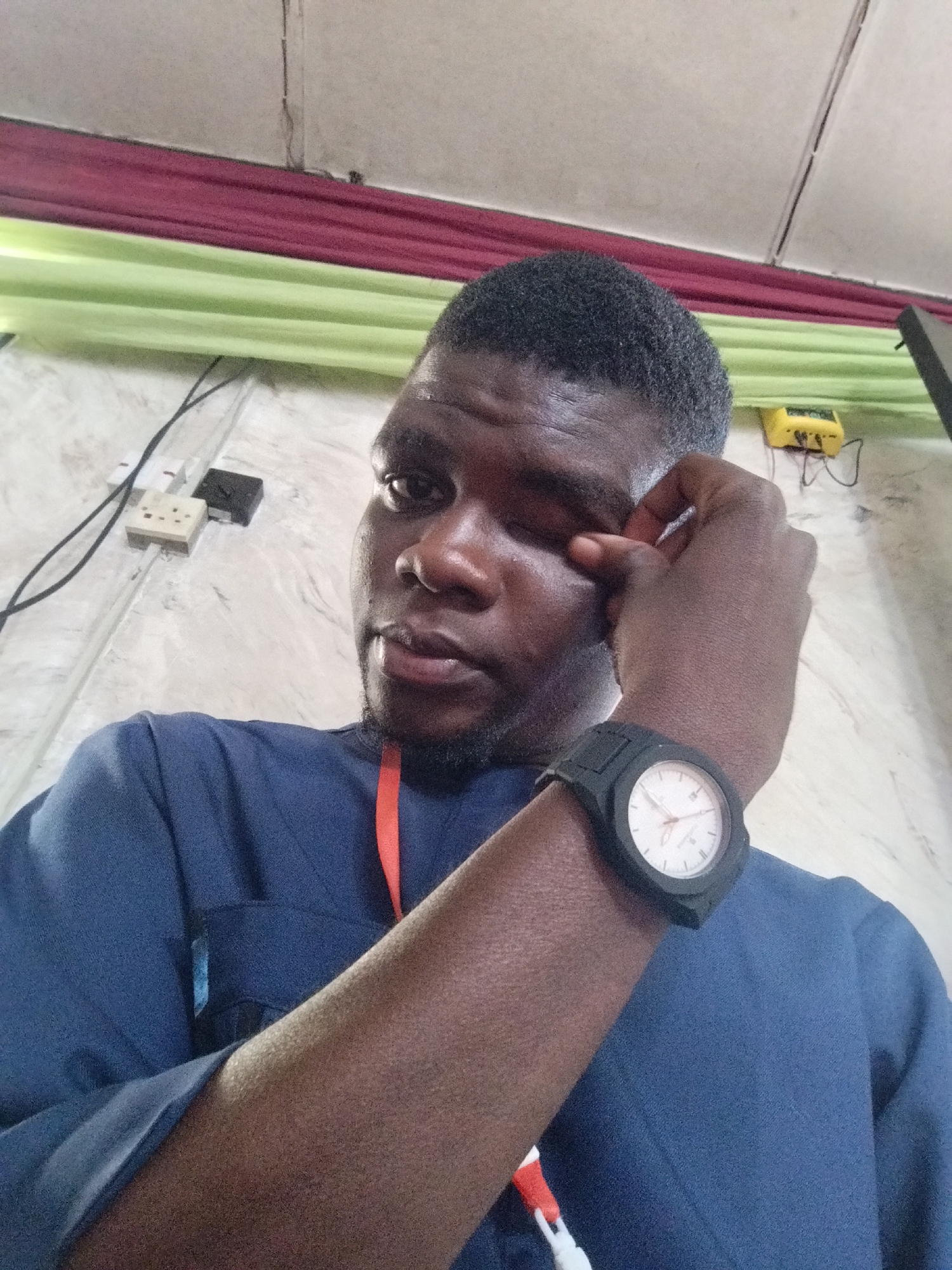 A person of African descent is photographed in an indoor setting. The individual, wearing a blue top, has a black watch on their left wrist and is touching their left eye with their left hand. Above the person are a wall with electrical outlets, a yellow electrical device, and a striped red and green decorative element near the ceiling. The location is noted as Unknown, Unknown.