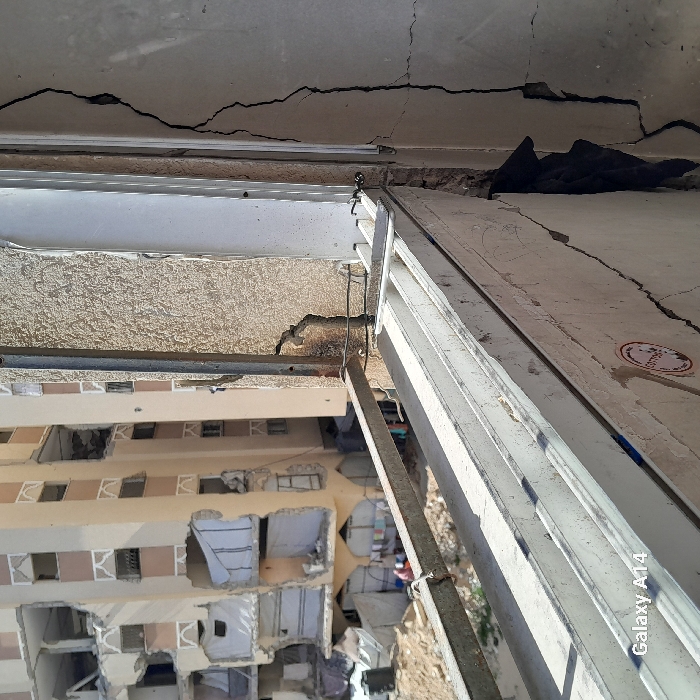 The image depicts a scene of extensive urban damage in the city of Khan Yunis, Palestinian Territory, viewed from an elevated position. In the immediate foreground on the right, a severely cracked, light-colored interior wall is visible, with a partially open or damaged white window frame. A vertical metal support bar is present, and a sticker with red elements is affixed to the lower wall. A rusty, horizontal metal railing extends across the lower midground.
Beyond the railing, a large, multi-story residential building, appearing light brown or beige, dominates the background. This building exhibits significant structural damage, with numerous sections of its facade missing, revealing interior rooms. Several exposed openings are covered with white tarpaulins, indicative of makeshift shelters or attempts to enclose spaces. At the ground level in front of this damaged building, multiple improvised shelters or tents are visible on sandy, disturbed earth. A black water tank is present near these shelters, and laundry is hanging outside one of them. Several individuals, including women and children, are discernable near the shelters, engaged in unidentifiable daily activities. The scene is illuminated by natural daylight.