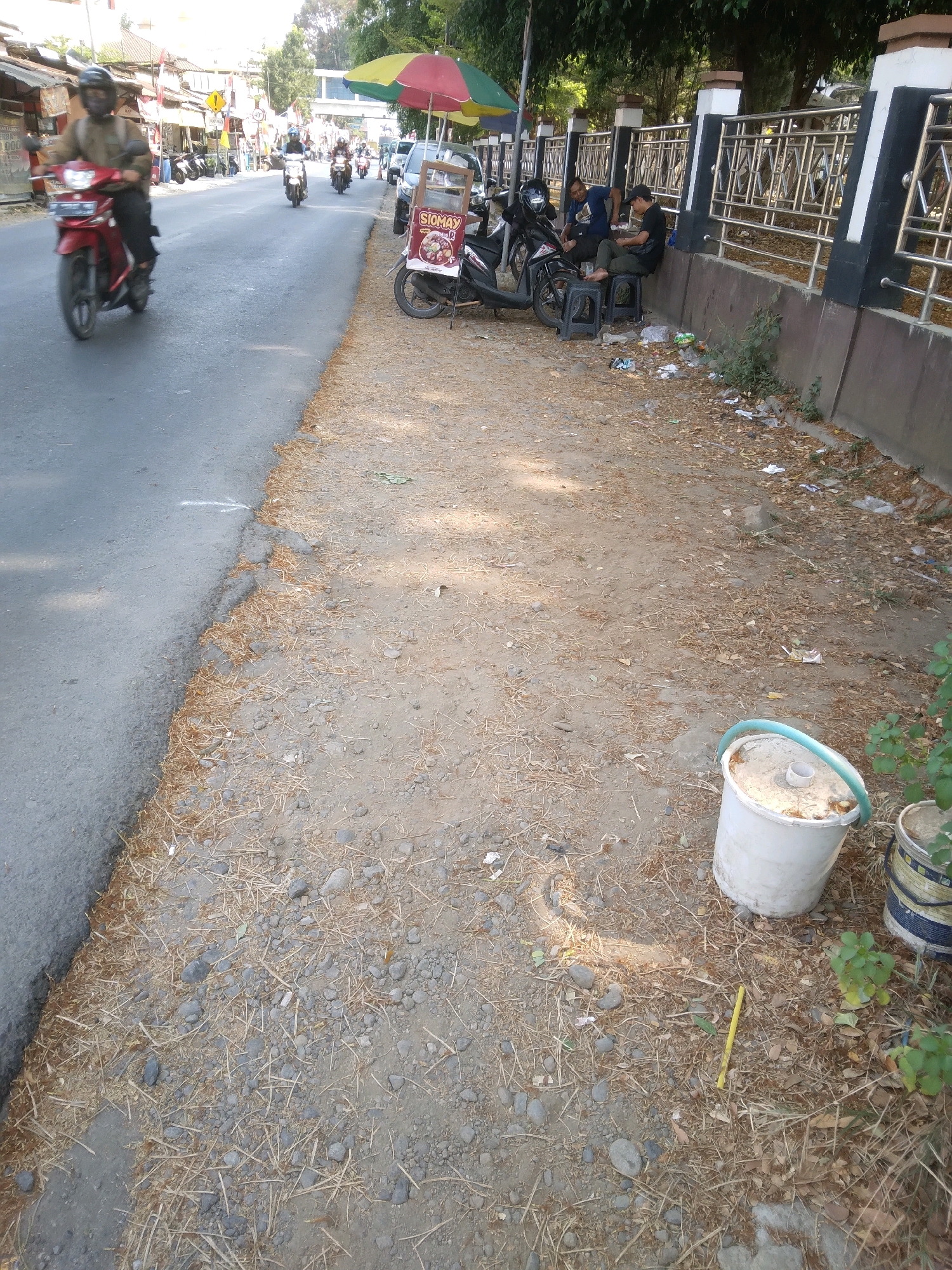 The image depicts a street scene in Desa Pamijen, Kecamatan Sokaraja, Indonesia. The foreground features a dirt and gravel roadside, littered with dry leaves, twigs, and scattered debris including plastic wrappers and bottles. A white bucket with a blue rim and a smaller white cylindrical object are present on the ground. A yellow stick lies nearby. The roadside transitions to a paved asphalt road where several motorcycles are visible. On the left, a red motorcycle is in motion, ridden by a person wearing a helmet and a brown jacket. Further down the road, other motorcycles are parked or in motion. On the right side of the road, a food stall with a colorful umbrella is set up, displaying a sign that reads "SIOMAY". Two individuals are seated on plastic stools near the stall, interacting with each other. Beyond the stall and roadside, a dark metal fence with decorative panels runs along a wall. Trees and foliage are visible in the background, along with buildings and a pedestrian overpass. The lighting suggests it is daytime, with sunlight casting shadows.