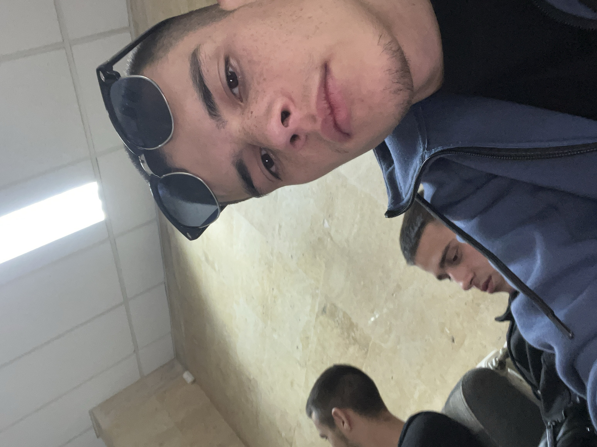 The image shows a selfie of a young man with short hair and sunglasses, wearing a blue and black jacket. Another person is partially visible behind him to the right, and a third person is seen from the back in the bottom center of the image. The background appears to be a room with tiled walls. The exact location in Plovdiv, Bulgaria, is not evident from the image.