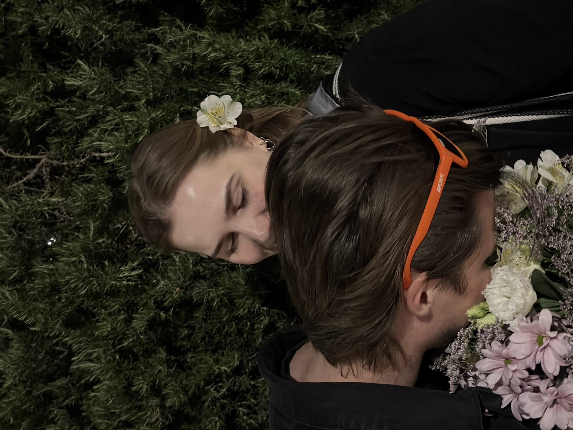 An angled, close-up shot captures two individuals amidst dense green foliage, likely at night or in low light conditions. A woman, positioned on the left side of the frame, has a white flower tucked into her light brown hair. Her eyes are closed, and she wears a silver hoop earring, conveying a sense of peace or rest.
To her right, a man with brown hair is seen in profile, his face turned towards a large bouquet of flowers he holds close, seemingly smelling them. He has light stubble and wears an orange headband or glasses pushed up on his head, with the text "APEROC" partially visible on the side. The bouquet features a variety of blooms, including prominent white and light pink/purple chrysanthemums or similar flowers, along with delicate sprigs that resemble dried lavender.
Both individuals are wearing dark clothing, the man a black jacket. The overall scene suggests a quiet, intimate moment, possibly outdoors, emphasizing sensory details like the scent of flowers and the serene atmosphere. The dark, bushy background provides a natural, somewhat secluded setting.