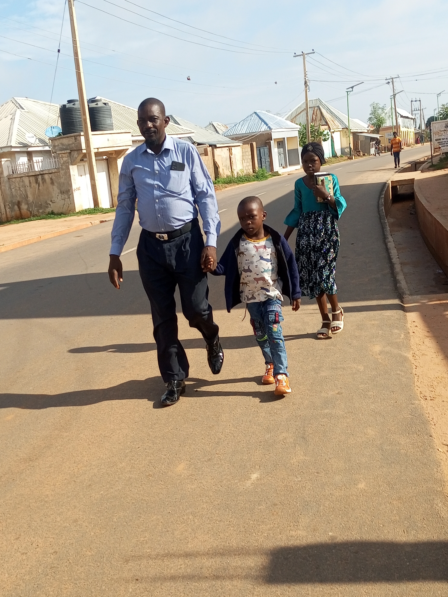 The image captures a sunny day in Gombe, Nigeria, likely on a street lined with modest buildings. In the foreground, a man, woman, and child are walking on the paved road, casting long shadows. The man, dressed in a blue striped shirt and dark trousers, holds the child's hand. He wears a black belt with a distinctive buckle and shiny black shoes. The child, a young boy, is wearing a t-shirt with a pattern of animals, blue jeans with patches, and orange sneakers. Behind them, the woman, who is wearing a teal top and a patterned skirt, also holds something in her hand. In the background, buildings with corrugated metal roofs are visible, along with utility poles and power lines. A few birds are in flight in the sky. To the far right of the image, a signpost with text is partially visible. The overall scene suggests a residential or commercial area within the city.