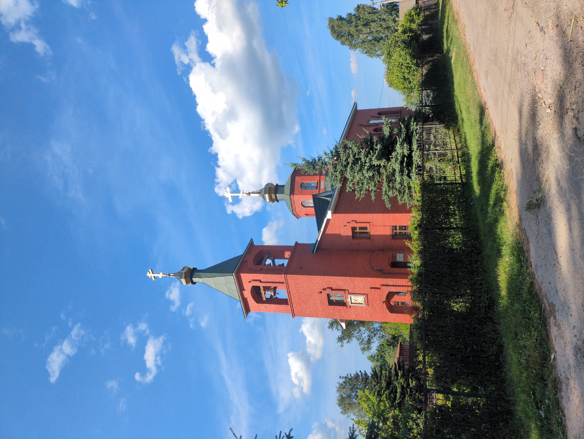 The image depicts an exterior view of a red brick Orthodox church with a green steeple and a golden cross, located in the city of Velet'ma, Russia. The church has a bell tower with two bells visible. The scene is set against a bright blue sky with scattered white clouds. In front of the church, there is a dark green hedge and a black metal fence. To the right of the church, there is a tall evergreen tree. The foreground shows a dirt road and some grass. The overall environment suggests a rural or suburban setting, bathed in sunlight on a clear day. There are no people visible in the image.