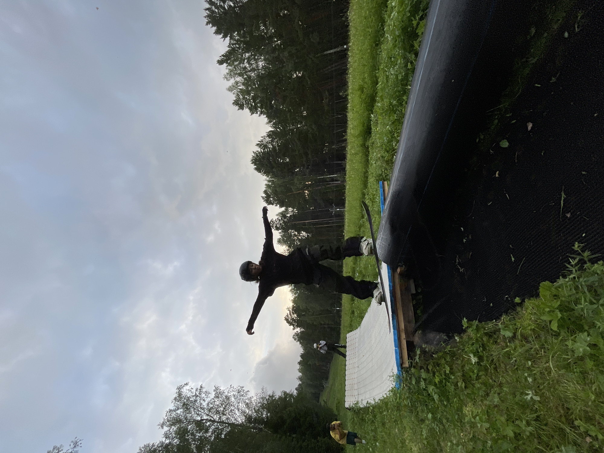 The image shows a person on a snowboard performing a trick in Divnogorsk, Russia. The person is wearing black snowboarding gear and a helmet, with arms outstretched as they balance on a snowboard. They are on a ramp covered with a white matting material, with a large black object, possibly a tunnel or obstacle, to their right. The scene is set outdoors in a grassy area with trees in the background, suggesting a snow park or a summer training facility. The sky is overcast and cloudy, indicating potentially cooler weather or an impending storm. The lighting suggests it is either early morning or late afternoon. There are other people visible in the background, some also on snowboards, and one person in a yellow shirt observing. The overall mood is one of outdoor activity and athletic pursuit.