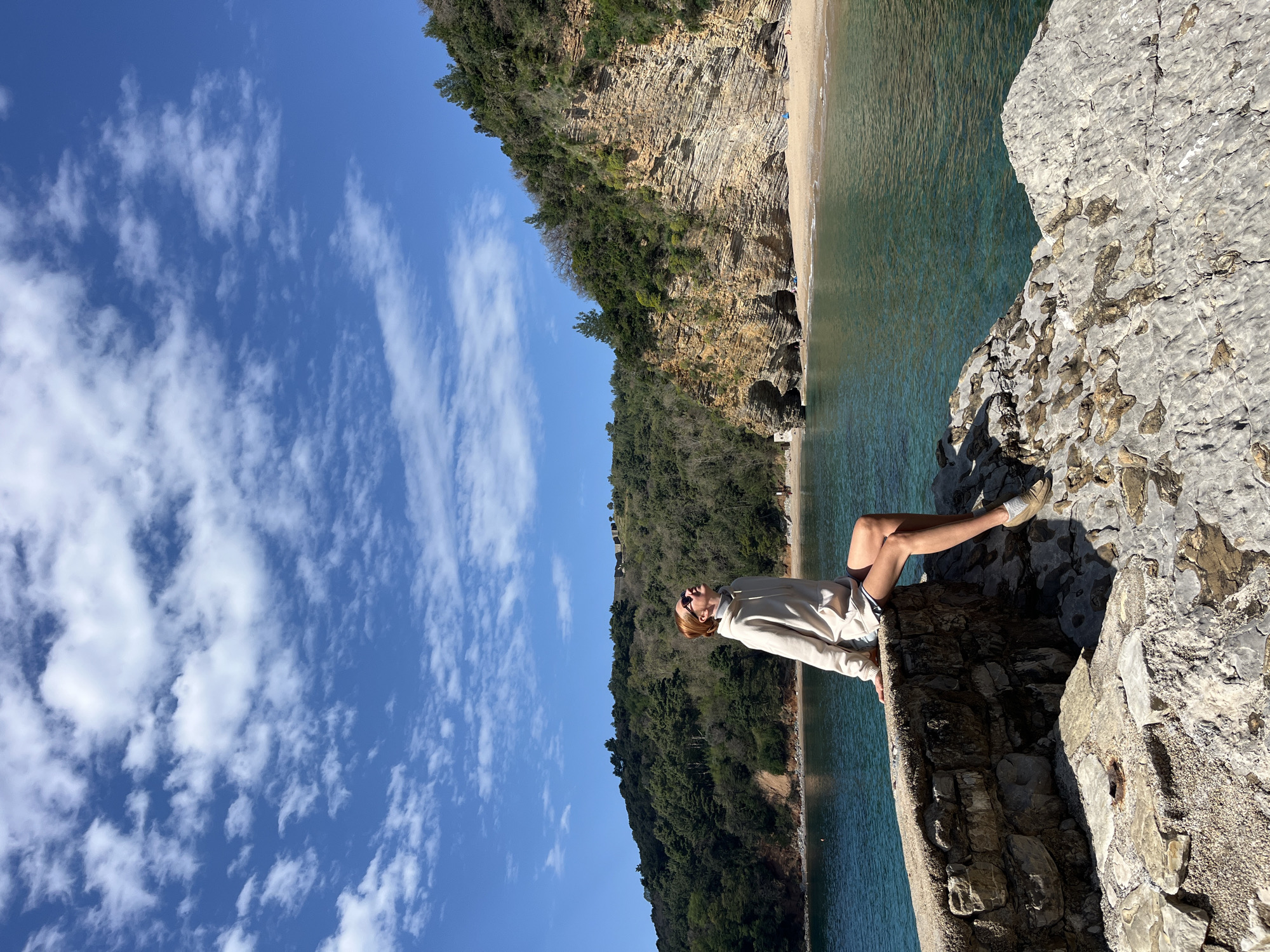 A medium outdoor shot depicts a person sitting on a rock wall overlooking a coastline. The person is wearing a light-colored hoodie and shorts with light-colored sneakers, and is resting against the wall. They are facing towards the coastline, which includes a sandy beach, turquoise water, and a cliff covered with greenery. In the background, there is a bright blue sky with scattered clouds. The location is likely in or near Budva, Montenegro, given the coastal landscape, rock formations, and vegetation characteristic of the area.