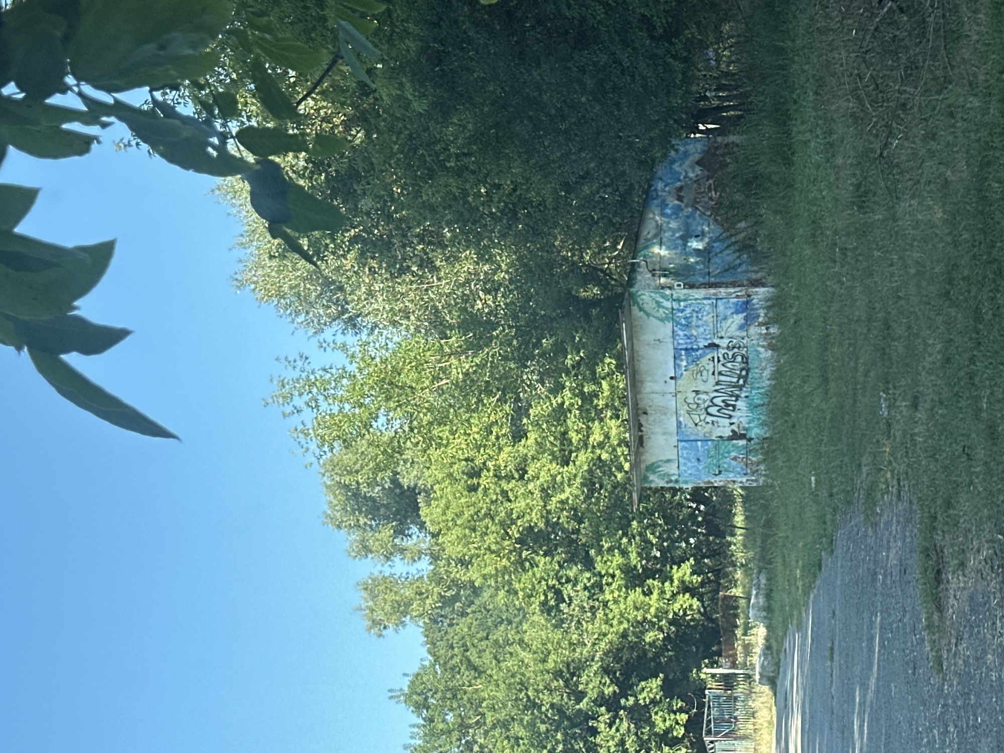 The image depicts a small, dilapidated building covered in graffiti, situated outdoors in what appears to be a rural or semi-urban setting. The building, with a corrugated metal roof and walls, is painted in various shades of blue, white, and green, with spray-painted tags and symbols. It is surrounded by overgrown grass and vegetation. In the background, a dense line of trees forms a backdrop, and a clear blue sky is visible above. To the left of the building, a paved path or road is visible, with a section of a colorful, metal fence in the distance. The overall scene conveys a sense of abandonment and neglect. The location in Makiv, Ukraine is not explicitly evident from the visual elements alone, but the context suggests an outdoor environment within the specified city.