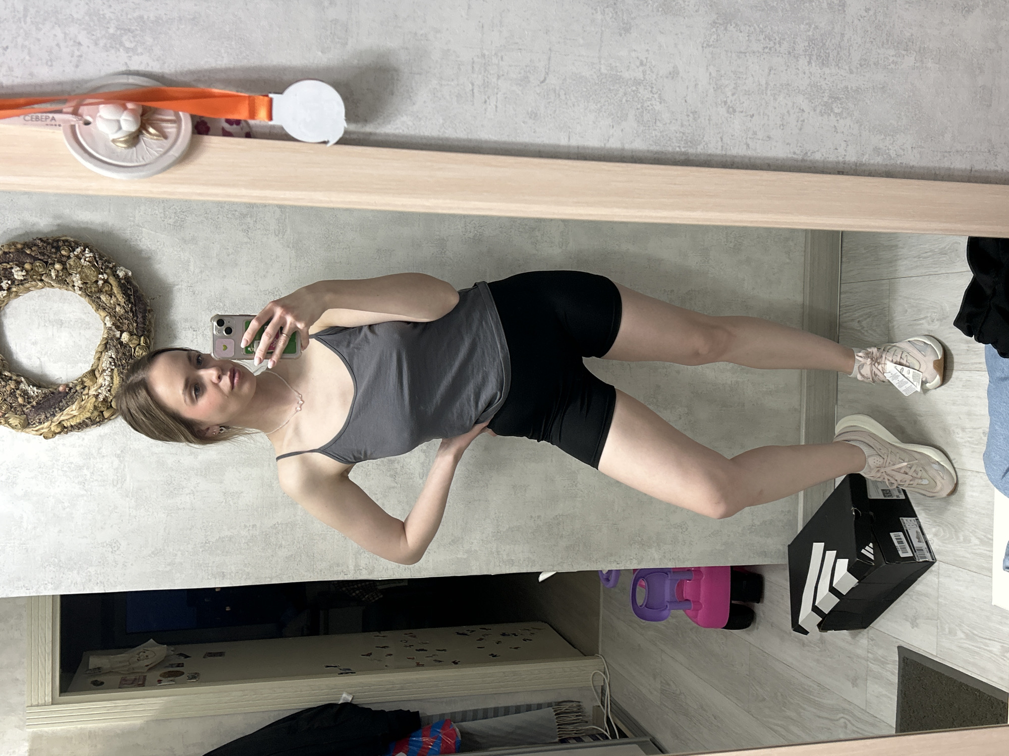 A young woman is taking a selfie in front of a mirror. She is wearing a gray tank top and black shorts, with pink sneakers on her feet. An Adidas shoebox is on the floor near her feet. The environment includes a wall with a wreath, a shelf with a medal hanging on it, and a view into another room revealing a TV, toys and personal belongings. The location is likely an apartment in Chaikovsky, Russia, based on the Cyrillic word "СЕВЕРА" visible on the medal.
