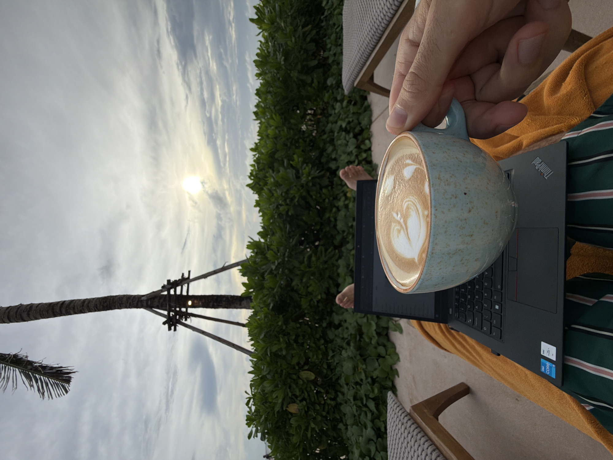 A view from a resort chair in Choeng Thale, Thailand, shows a person's hand holding a light blue, speckled ceramic mug of latte art over a grey ThinkPad laptop on a lap covered with striped fabric and a tan towel. A dark green hedge runs horizontally through the mid-ground. A palm tree stands to the left, its trunk supported by wooden beams. The sky is grey with the sun breaking through the clouds.