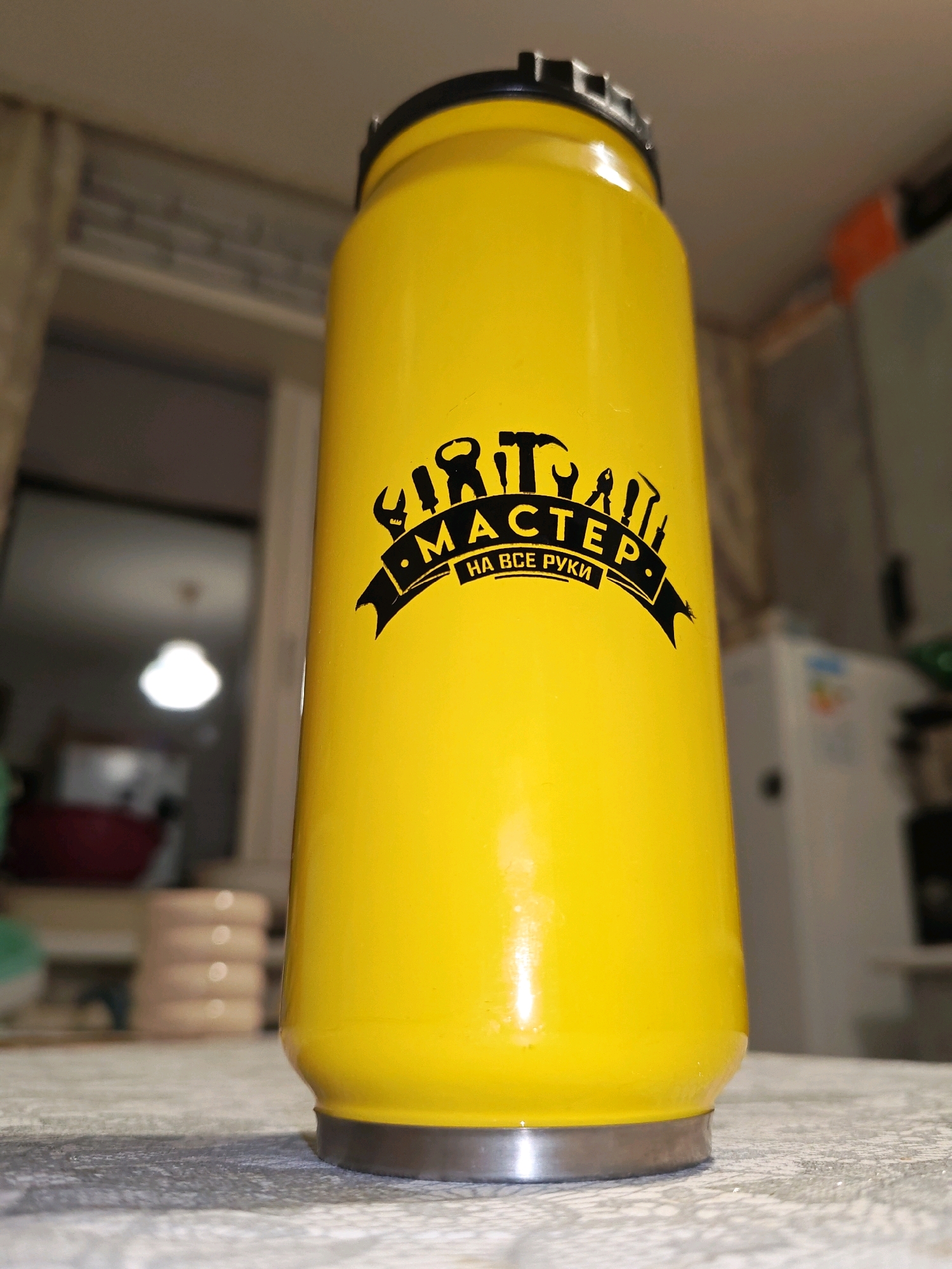 A low-angle, close-up shot shows a bright yellow, cylindrical object with a black lid, positioned on a textured surface. The object features a black graphic logo consisting of various tools such as a hammer, wrench, and screwdriver, arranged above text that reads "МАСТЕР НА ВСЕ РУКИ." The background is blurred, revealing indoor elements like curtains, a light fixture, and kitchen appliances, suggesting an indoor setting, possibly a kitchen or a workshop. The object appears to be a thermos or a similar container. The image is not taken in the city of Chaikovsky, Russia; this is a misattribution.