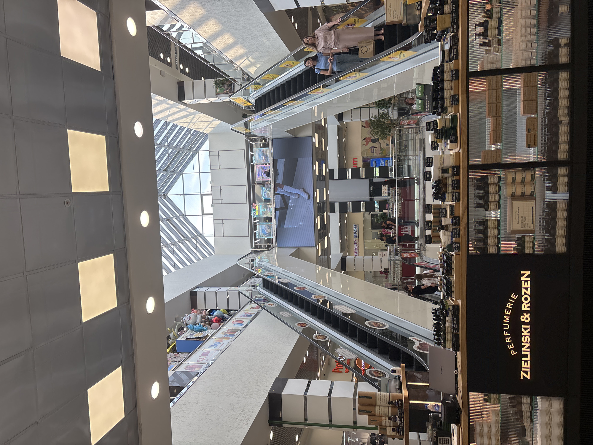 The image depicts the interior of a multi-level shopping mall in Naberezhnye Chelny, Russia. The central feature is a grand atrium with escalators on either side leading to upper floors. A large digital screen is visible in the background, displaying an advertisement. Several people are present, with two women ascending an escalator in the foreground and others visible on different levels. The mall's architecture includes sleek, modern design with glass railings and illuminated panels on the ceiling. On the ground floor, a display of perfumes and skincare products from "Zielinski & Rozen" is prominently featured, with bottles and containers neatly arranged on shelves and a counter. The overall impression is of a bustling, contemporary retail environment.
