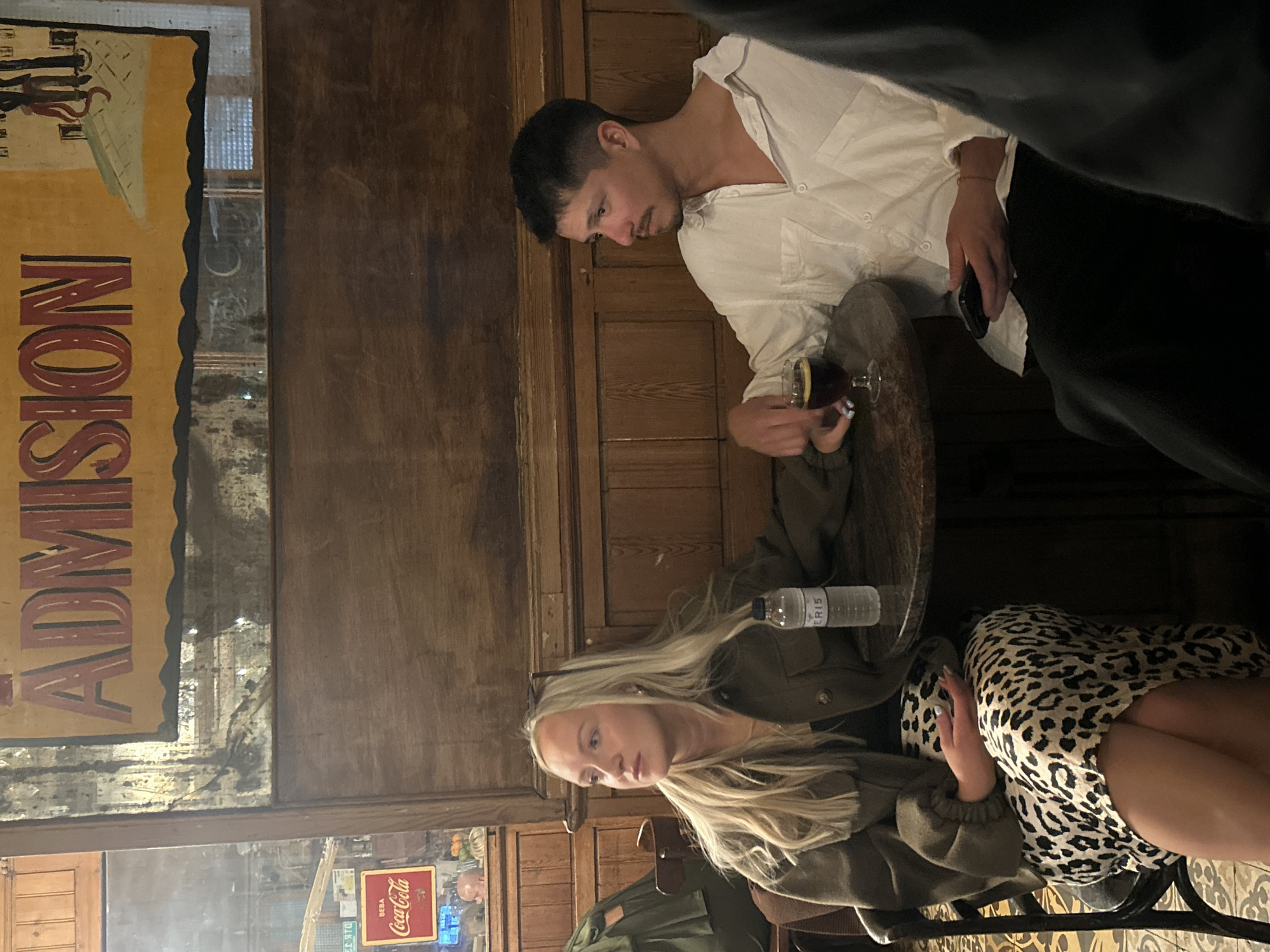 Two individuals are seated at a table within a dimly lit establishment, possibly a bar or restaurant. A fair-skinned female with long blonde hair, wearing a green jacket and a leopard print dress, is seated on a chair and appears to be looking at something or someone in the middle distance. Next to her, a male with short dark hair is standing and holding a glass with a lemon in it. He is wearing a white shirt and dark trousers. A mobile phone is in his left hand. A water bottle sits on the table. The environment is marked by dark wood panelling and an old-fashioned Coca-Cola sign that can be seen through the window, suggesting a vintage interior. A sign with the word "ADMISSION" is visible on the left. Based on the style and architecture, and the "Coca-Cola" sign written in the Catalan language ("BEBA"), the location appears to be a historical building in Barcelona, Spain.