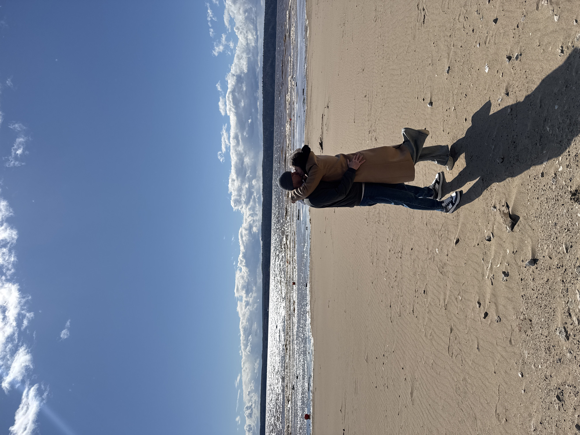 An outdoor scene captures a romantic moment between two individuals on a sandy beach under a bright blue sky. The image is oriented with the sky at the top, a body of water in the middle, and sand at the bottom.
In the foreground, a couple is embracing and kissing. The person on the left, wearing a dark jacket, blue jeans, and a grey beanie, holds the person on the right, who is wearing a long beige coat and dark pants. Their long, dark shadows stretch out behind them onto the sand, indicating a clear, sunny day, likely late morning or early afternoon. The sand beneath them is light brown, showing ripples and some scattered small stones and debris.
Beyond the couple, a wide expanse of water sparkles intensely under the sun, reflecting bright highlights across its surface. In the distance across the water, a dark, narrow strip of land covered with trees or foliage is visible, forming the opposite shore. The sky is a clear, vibrant blue with scattered white, fluffy clouds, particularly along the left side.
The overall atmosphere suggests a serene and affectionate moment amidst natural beauty. There is no visible text in the image. Given the location context of Polazna, Russia, the body of water is likely the Kama Reservoir.