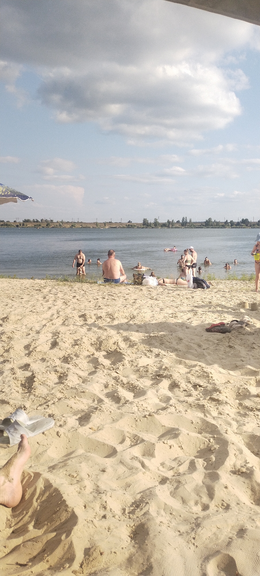 The image depicts a sandy beach next to a body of water, with several people engaged in various activities. The scene is set in Bezliudivka, Ukraine, as indicated by the context.
In the foreground, a person's foot wearing a sandal is visible, resting on the sand. Further into the sand, there are scattered items like more sandals and a bag.
The middle ground features several individuals on the beach and in the water. Some are lying on the sand, sunbathing, while others are standing or wading in the water. A man is seated on the sand facing the water, with his back to the camera. A larger group of people are in the water, some swimming or floating. An umbrella is partially visible in the upper left corner.
The background shows a calm body of water, beyond which is a tree line and a distant, faint structure that could be power lines or a distant building. The sky is partly cloudy, with a mix of blue sky and white clouds. The overall impression is a leisurely day at the beach.