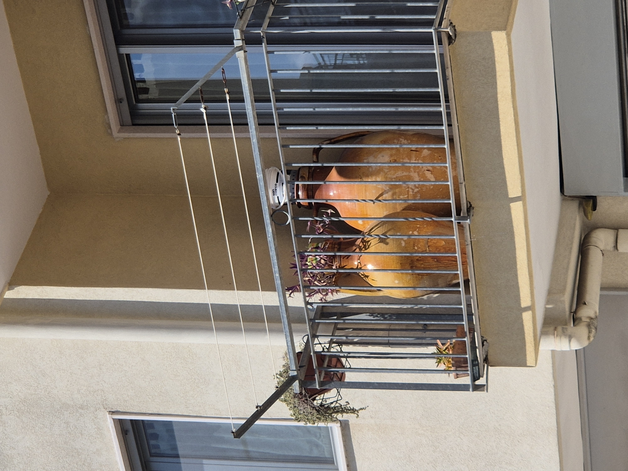 The image shows a balcony on the exterior of a building in Laterza, Italy. The balcony is made of metal railings and has a clothesline strung across it. There are two large, orange ceramic jars on the balcony, one of which has a white container on top with the text "san marco". In the background, there are windows and part of the building's facade. There are also plants in pots on the balcony. The lighting suggests it is daytime, with sunlight casting shadows.