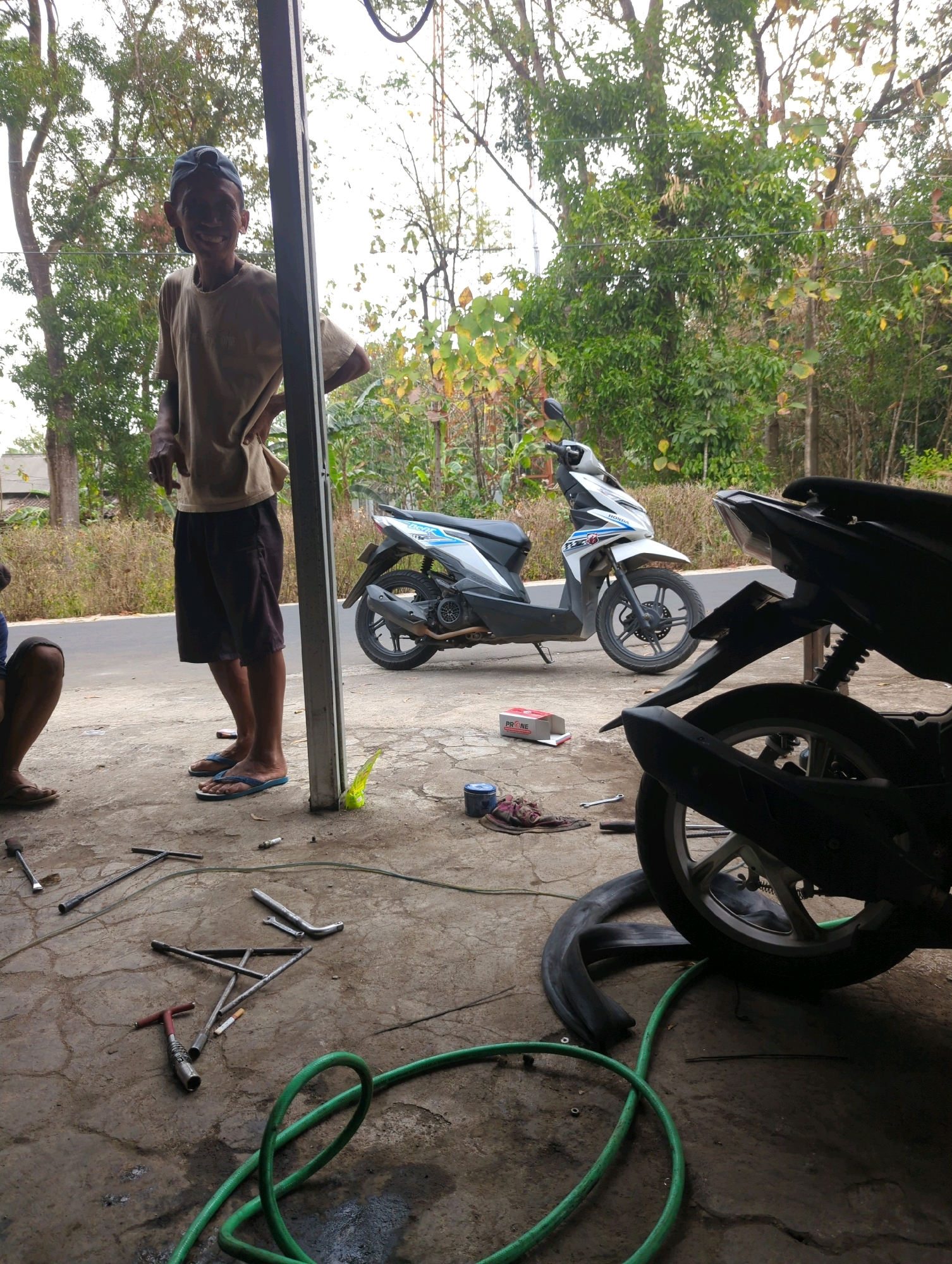 A man wearing a light brown t-shirt and dark shorts stands with his hands on his hips, looking at the camera. He is wearing a blue baseball cap. In the background, two motorcycles are parked. The ground is paved with concrete and covered with tools, including wrenches and a tire tube. A green hose lies coiled on the ground. The scene appears to be an outdoor workshop or garage in Kedungdowo, Indonesia.