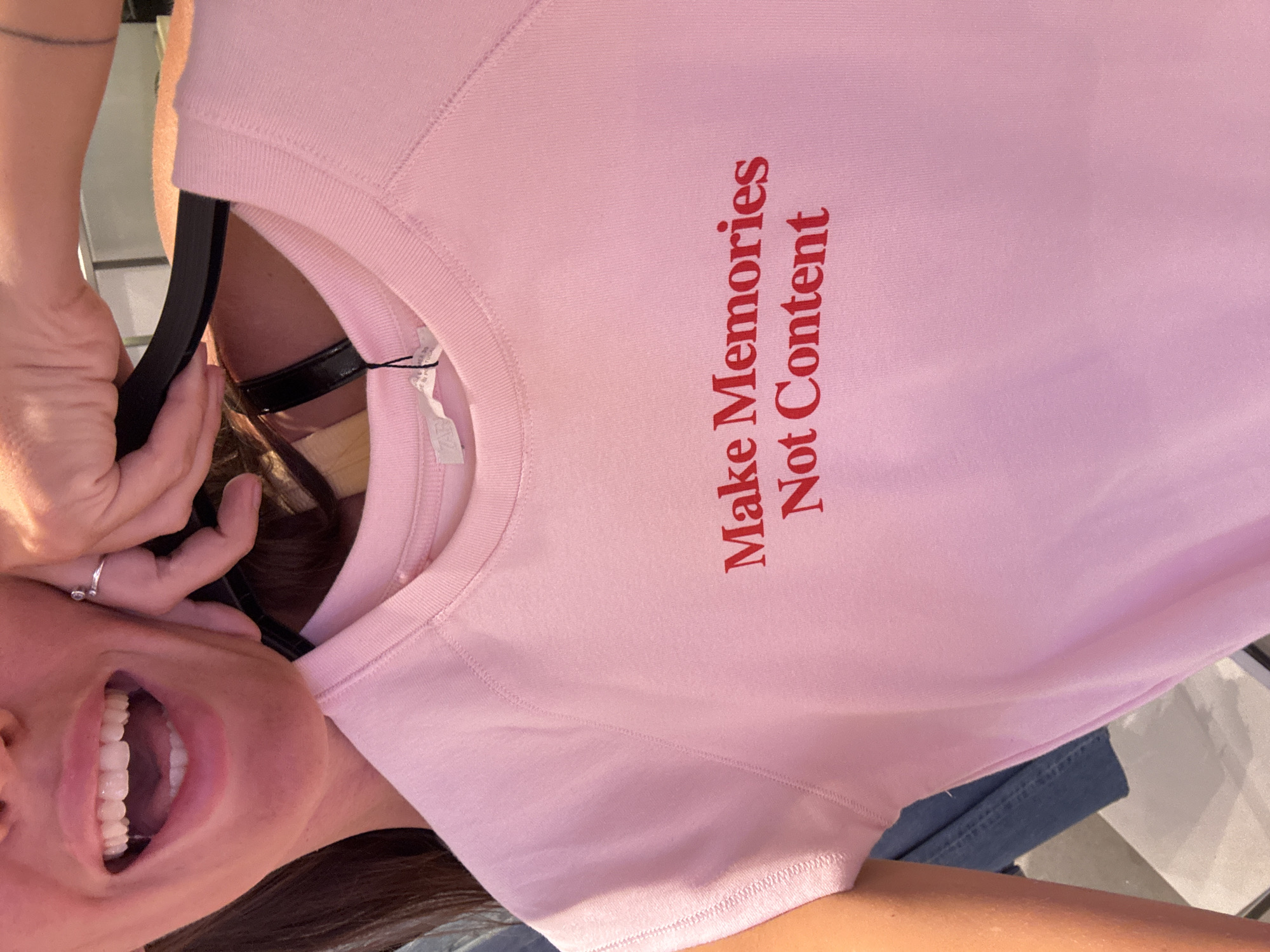 A woman with long brown hair is holding a light pink sweatshirt on a black hanger in front of her. The sweatshirt has red text that says "Make Memories Not Content". She is smiling broadly, showing her teeth. The background is blurry, but suggests an indoor location with light-colored surfaces. She is wearing blue jeans. The location is not possible to determine based on the image provided.