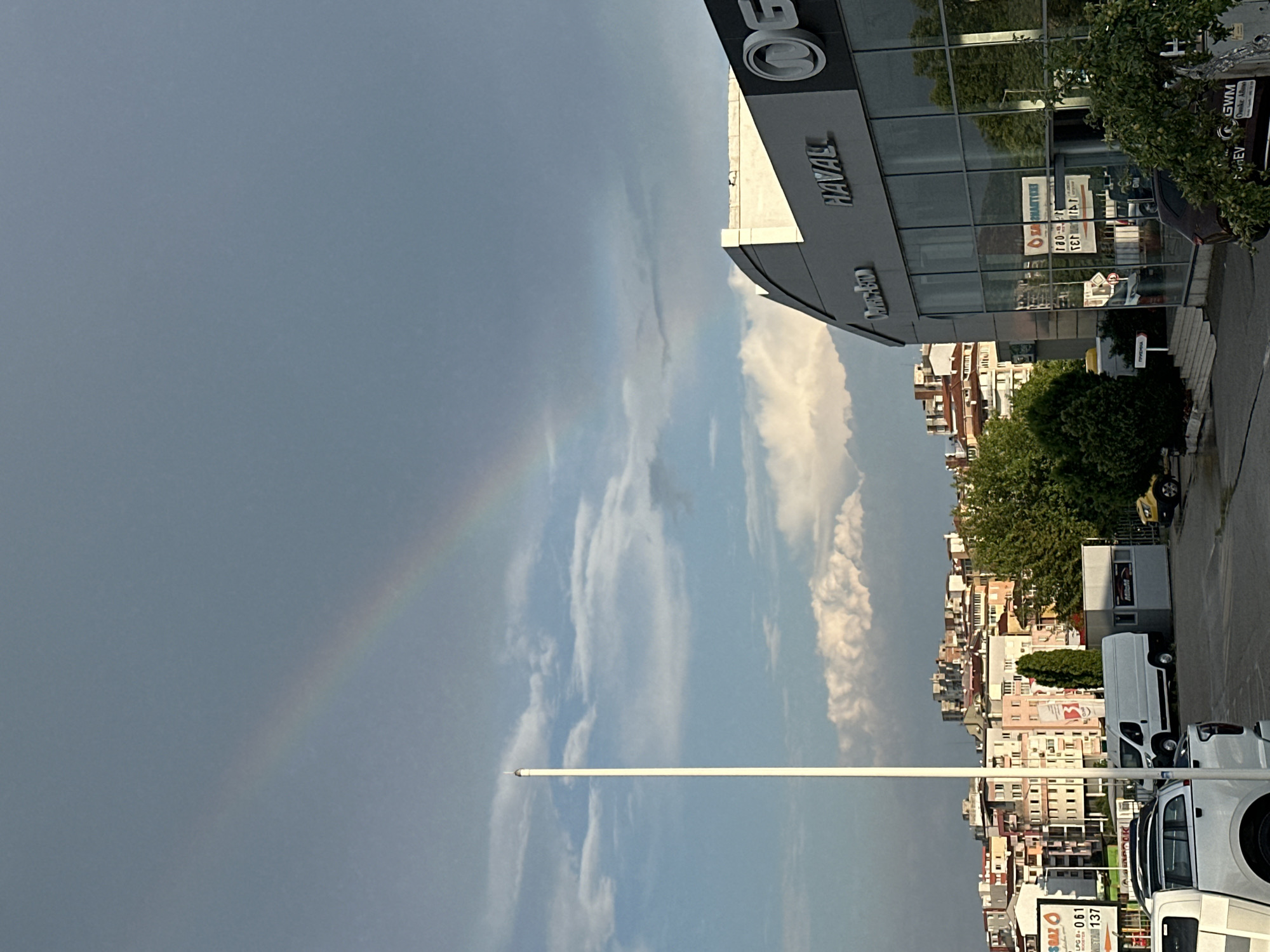 The image depicts a street scene in Veliko Tarnovo, Bulgaria, under a dynamic sky. A faint rainbow arcs across the left side of the frame, suggesting recent rain or an ongoing shower. The sky is a mix of soft grey clouds and brighter patches of blue, indicating a changeable weather pattern, likely late afternoon or early evening given the light.
Dominating the right side of the image is a modern building with a glass facade, likely a car dealership, as evidenced by the prominent "HAVAL" logo and other signage visible through the windows. Reflections of greenery and buildings are visible on the glass. A white van and part of a white pickup truck are parked on the street in front of the building.
Behind the dealership, a dense urban landscape unfolds, characterized by multi-story buildings with varied architectural styles and colors. Trees are scattered throughout the urban environment, adding touches of green against the backdrop of the city. A tall, white pole extends vertically through the center of the frame, its purpose unclear.
The overall impression is one of a moment captured during a transition in weather, with the urban setting of Veliko Tarnovo providing a detailed backdrop. Visible text includes "HAVAL", "GWM", "NAVAL", and partial signs with Cyrillic lettering such as "ПРЪРЖЕНА" and "SMART".
