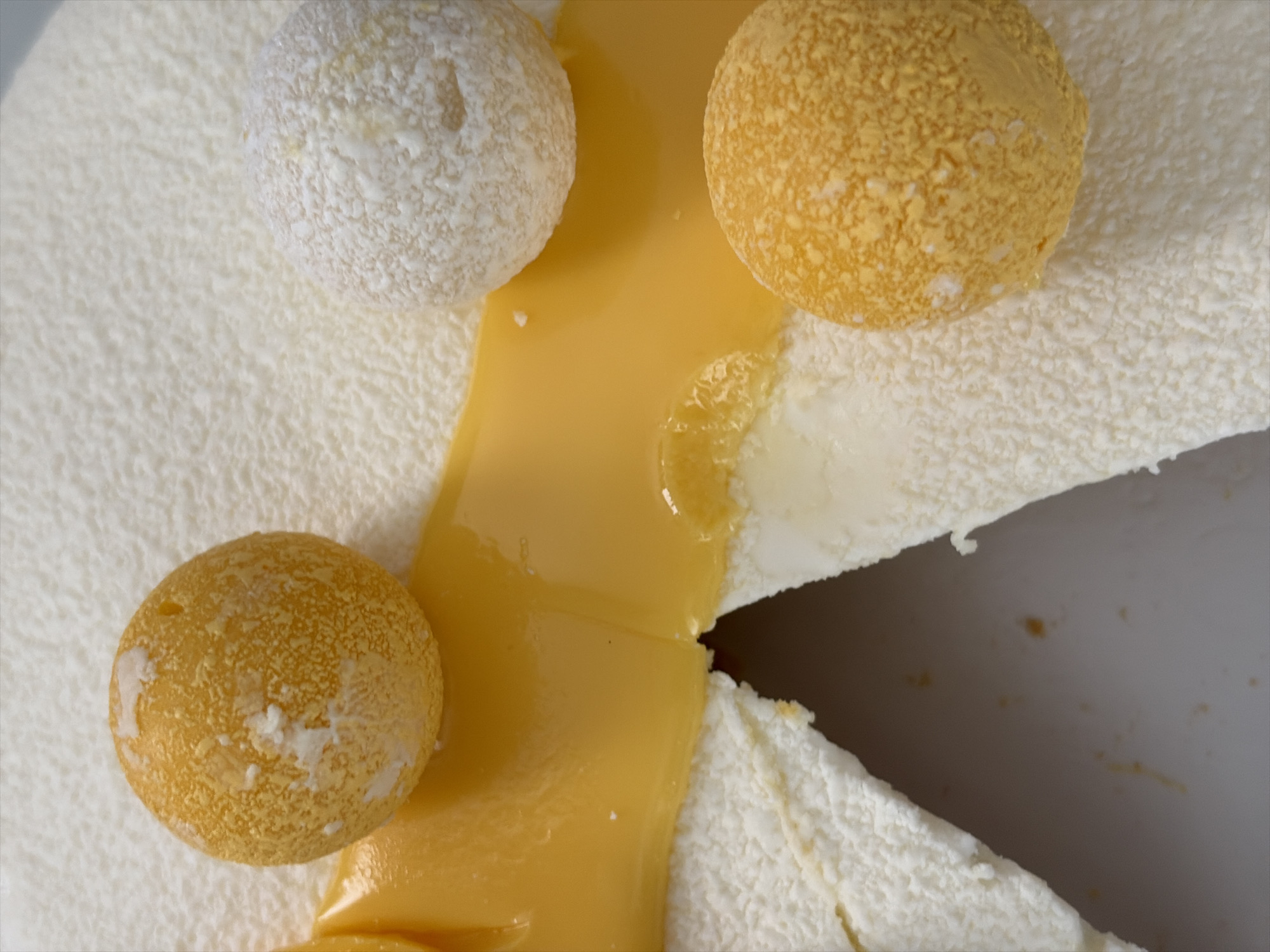 The image is a close-up, overhead view of a dessert. The dessert features a white, textured outer layer, possibly cake or mousse, with cuts revealing a creamy interior. A vibrant yellow, translucent gel or sauce is spread across the surface. Several spherical toppings are visible: two are a bright orange-yellow with a textured, slightly crumbly surface, and one is white, also with a textured appearance. One orange sphere is positioned on the left, partially submerged in the yellow sauce. Another orange sphere is on the lower right, resting on the white outer layer. The white sphere is located on the upper right, on the yellow sauce. The background is blurred and appears to be a light-colored surface, possibly a plate or table. There are no identifiable landmarks or context to suggest a location in Moscow, Russia.