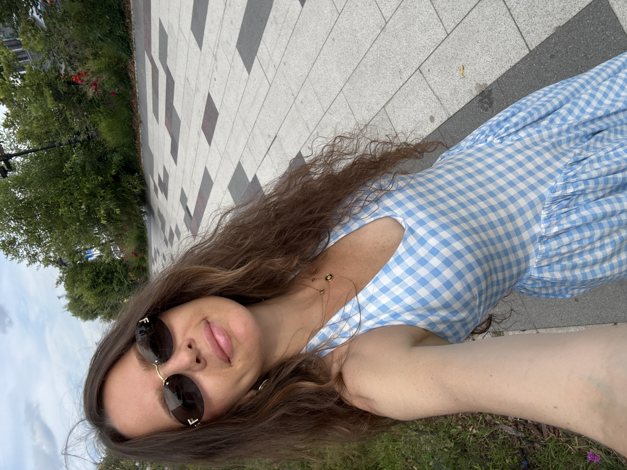 A woman with long, wavy brown hair is taking a selfie outdoors. She is wearing a blue and white gingham sundress, sunglasses with prominent "F" logos, and a delicate gold necklace. The woman has light skin and her lips are slightly parted. She appears to be smiling faintly.
The background suggests a park or garden setting with a paved walkway made of rectangular tiles in alternating grey and white patterns, and patches of green grass and trees. Some red flowers are visible among the foliage. The sky is overcast, indicating a cloudy day. The orientation of the photo suggests it may have been taken around midday. The visible details like the paving stones and greenery are consistent with an urban outdoor space in Moscow, Russia. There is no visible text in the image.