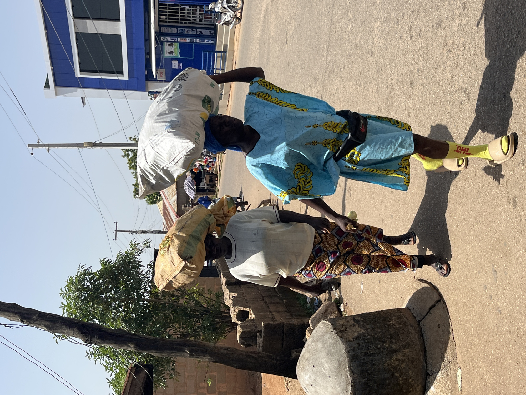 The image shows two individuals carrying large sacks of goods on their heads. The person in the foreground, on the right, is wearing a blue patterned dress and yellow socks with "DHL" printed on them. They also have a black fanny pack around their waist. The second person, on the left, is wearing a white shirt and a colorful patterned skirt. Both are walking on a paved street in what appears to be a town or city.
Behind them, a blue and white building with large windows is visible, possibly a shop or business. Power lines crisscross the sky, suggesting an urban or semi-urban environment. A tree with green leaves is also present in the scene. The lighting suggests it is daytime, with strong shadows indicating a sunny day. The overall atmosphere appears to be one of daily life and commerce, with people engaged in the task of transporting goods. There is no visible text other than the "DHL" on the socks.