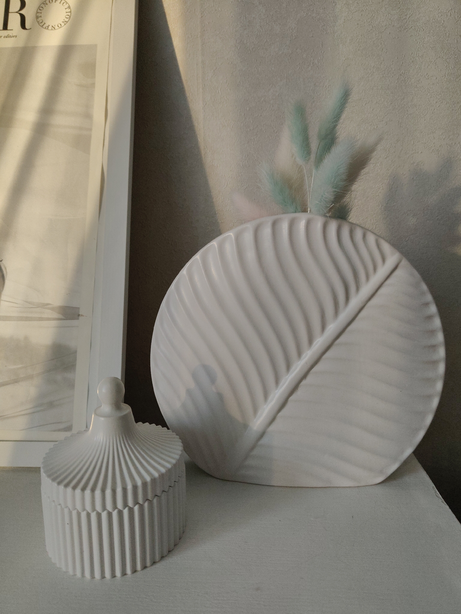 The image depicts a still life arrangement of decorative objects. On a white surface, likely a shelf or table, are two prominent items: a white, round vase with a leaf-like embossed design and a small, white decorative jar with a lid, featuring a ridged pattern. The vase contains a few sprigs of light blue or green ornamental grasses. Adjacent to the vase is a framed picture with visible text. The environment appears to be an interior space, judging from the lighting and the presence of furniture. The location is not specified, rendering the city and region Unknown, Unknown.