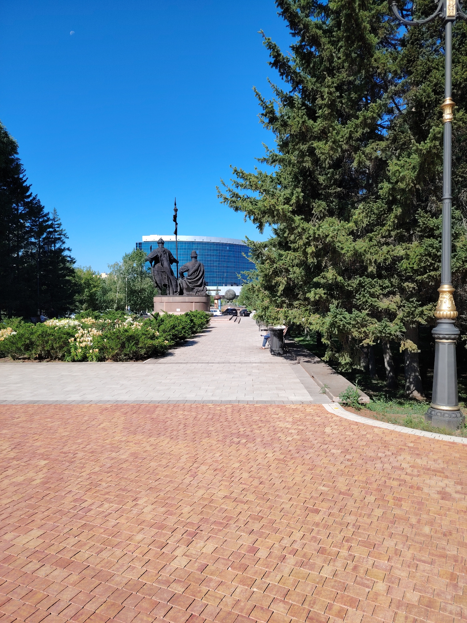 A sunny day in Astana, Kazakhstan, features a wide, brick-paved walkway in the foreground that curves to the left. Beyond the walkway, a paved path leads towards a statue of two figures, a man standing and another kneeling, positioned on a pedestal. In front of the statue is a bed of white and yellow flowers bordered by lush green bushes. To the right of the path, a large evergreen tree shades a bench where a person is seated, looking away from the camera. Behind the statue and flowers, a modern, glass-fronted building with a blue facade stands tall. To the far right of the image, a decorative lamppost with gold accents is visible. The sky is a clear, vibrant blue with a sliver of the moon visible in the upper left corner.