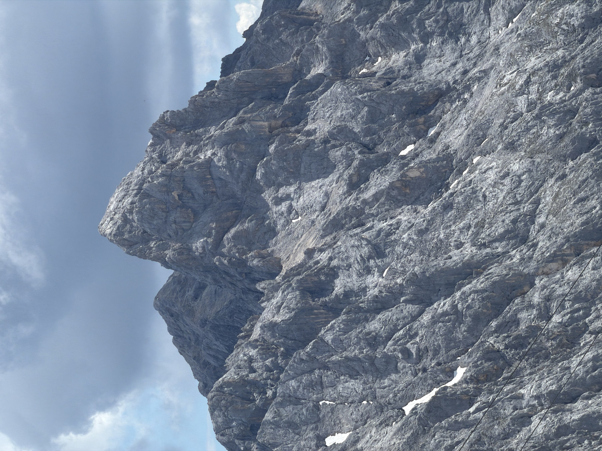 The image is a close-up, eye-level, medium shot of a rocky mountain peak under a cloudy sky. The mountain is composed of grey, jagged rock formations with some patches of snow scattered across its slopes. The sky is predominantly overcast with dramatic, dark grey clouds, but hints of blue sky are visible in the upper left corner. The overall impression is one of rugged, natural grandeur. This mountain is Zugspitze, the highest peak in Germany, located in the Bavarian Alps, bordering Austria, and is a prominent natural landmark near Garmisch-Partenkirchen. There are no people or man-made objects visible in the image.