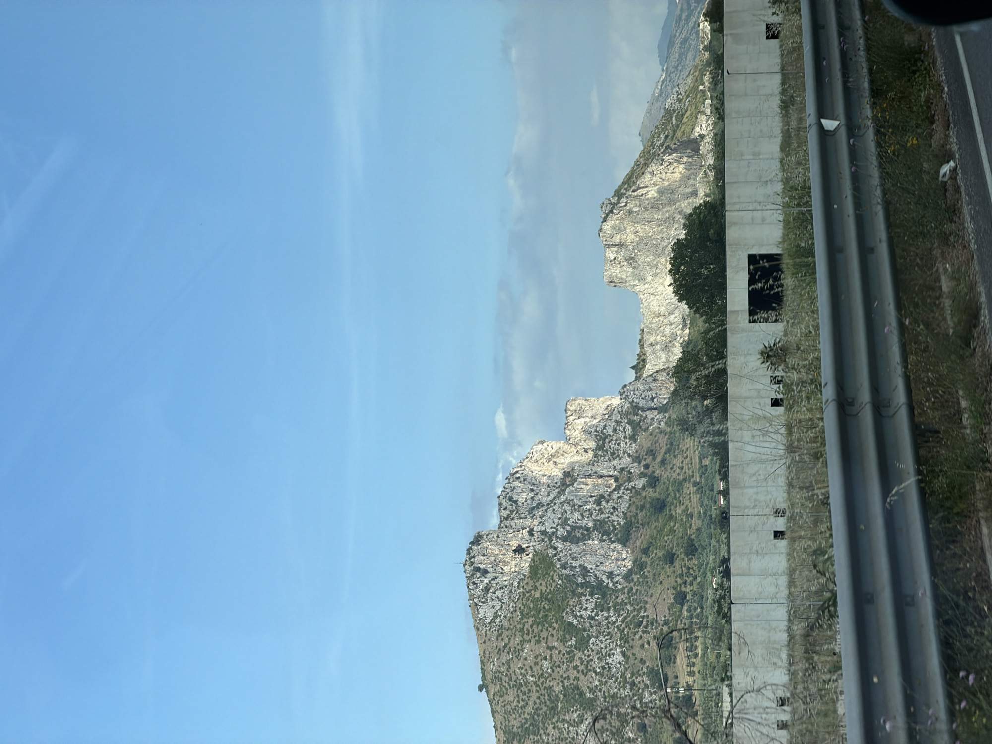 This portrait-oriented photograph captures a roadside landscape scene under a bright sky, seemingly taken from inside a moving vehicle.
The left side of the image is dominated by a clear, vibrant blue sky, with subtle, wispy white clouds visible, particularly towards the center. A range of rugged mountains stretches across the mid-ground, characterized by their rocky, light-colored faces interspersed with areas of sparse green and brown vegetation, suggesting a dry, Mediterranean climate. Sunlight brightly illuminates parts of the mountain peaks and slopes.
The right side of the image features elements of a highway. A grey metal guardrail runs vertically along the right edge, separating the roadway from a grassy verge. The verge contains dry grasses, some small flowering plants with purple blooms, and a discarded white plastic bag near the road surface. Behind the guardrail, a tall, light-colored concrete retaining wall or sound barrier extends along the road. This wall has several evenly spaced, rectangular cutouts. Beyond this barrier, more trees and the lower slopes of the mountains are visible.
The scene depicts a sunny day, likely late morning or early afternoon, given the intensity and angle of the light. There are no people visible, nor is there any discernible text. The image conveys a sense of travel through a mountainous region, aligning with the location context of Santa Maria Capua Vetere, Italy.