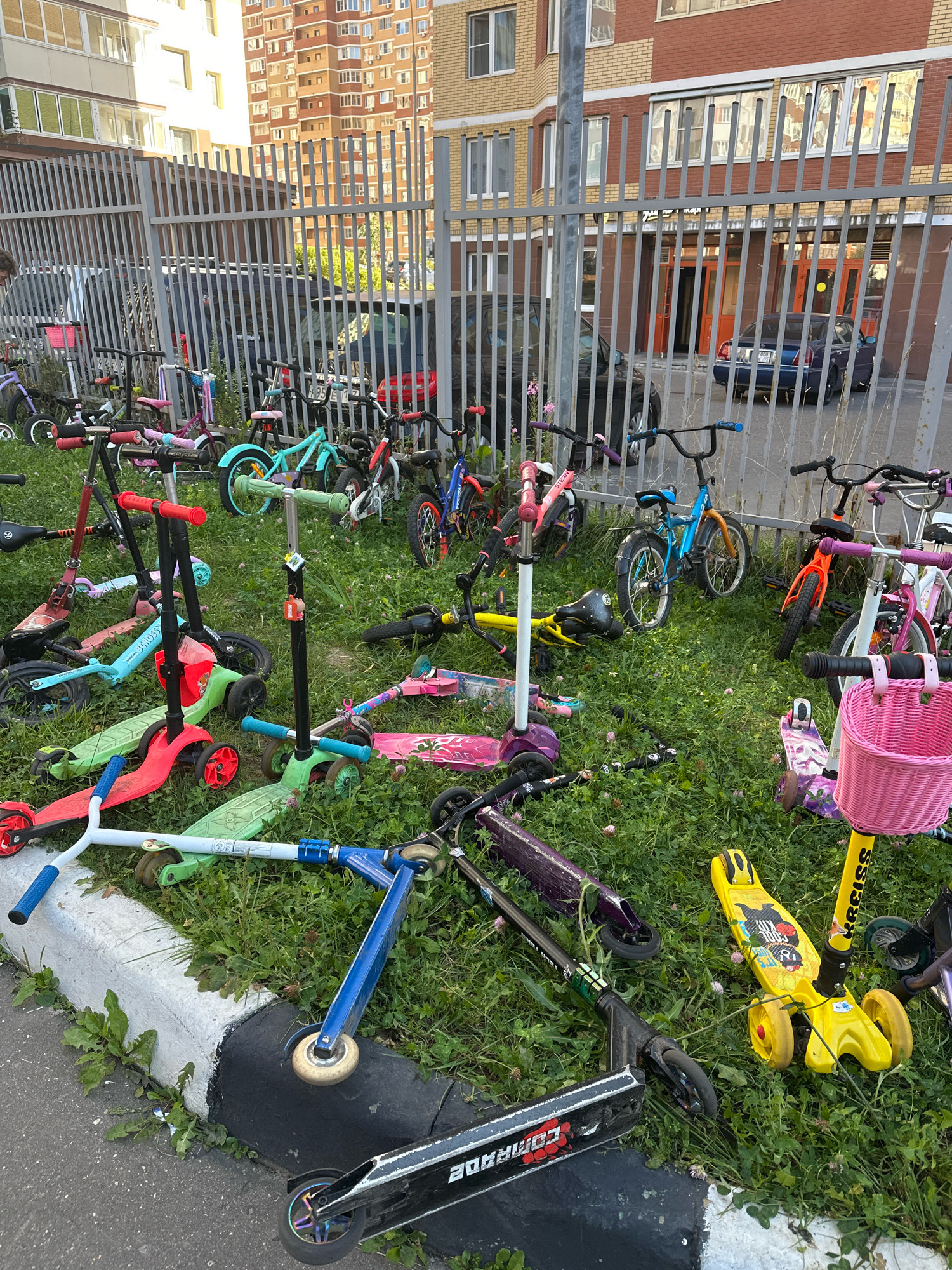 A street view of children's scooters and bicycles are scattered on the grass in front of a metal fence. A blue sedan is parked behind the fence. In the background, multi-story apartment buildings are visible. This scene is located in Vidnoye, Russia.