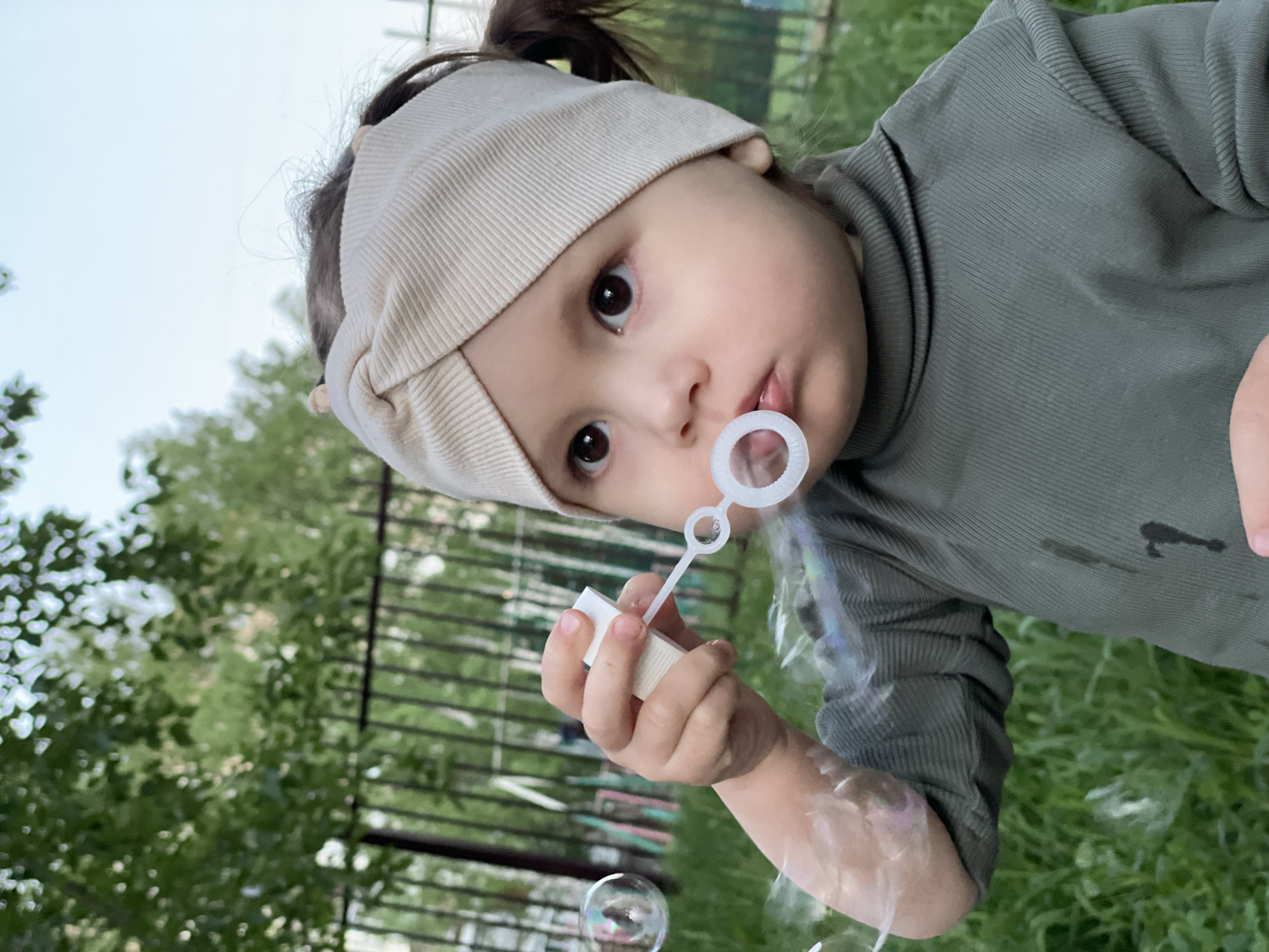An outdoor, eye-level candid portrait captures a young child, likely a toddler or preschooler, in Izhevsk, Russia, actively blowing bubbles. The child, looking directly at the camera with wide, dark eyes and a slightly open mouth, holds a white plastic bubble wand close to their lips. They are wearing a light beige ribbed headband and a dark olive-green ribbed long-sleeved shirt.
Numerous iridescent bubbles float around the child, reflecting light. The background features vibrant green foliage, indicative of trees or bushes, and a dark metal fence. The ground visible in the lower part of the frame is green grass. The sky above is bright, suggesting daytime with soft, possibly overcast, lighting. The overall image has a tilted orientation, adding to its spontaneous feel, and the child's expression conveys curiosity and engagement with the activity. No visible text is present in the image.