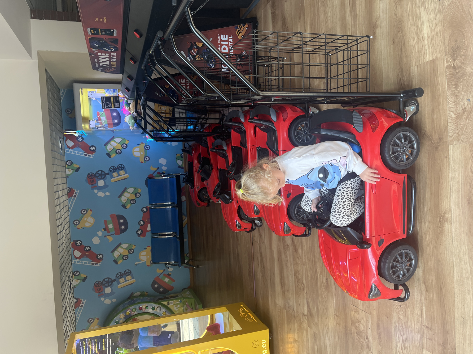 In an indoor setting, a young girl with blonde hair, wearing a white sweatshirt with a blue character and patterned leggings, sits in a red toy car. The car has a black interior and black wheels. Behind her, several other identical red toy cars are lined up, some with black interiors and wheels, and others with just the car bodies visible. To the right, black metal carts with wire mesh compartments are positioned next to the cars. In the background, blue benches are visible, and a wall is decorated with a playful wallpaper featuring various cartoon vehicles like boats, cars, and trucks. Further back, arcade machines are present, one with a yellow frame displaying text, and another red machine with the text "Kiddie Cart Rental". The floor is made of polished wood.