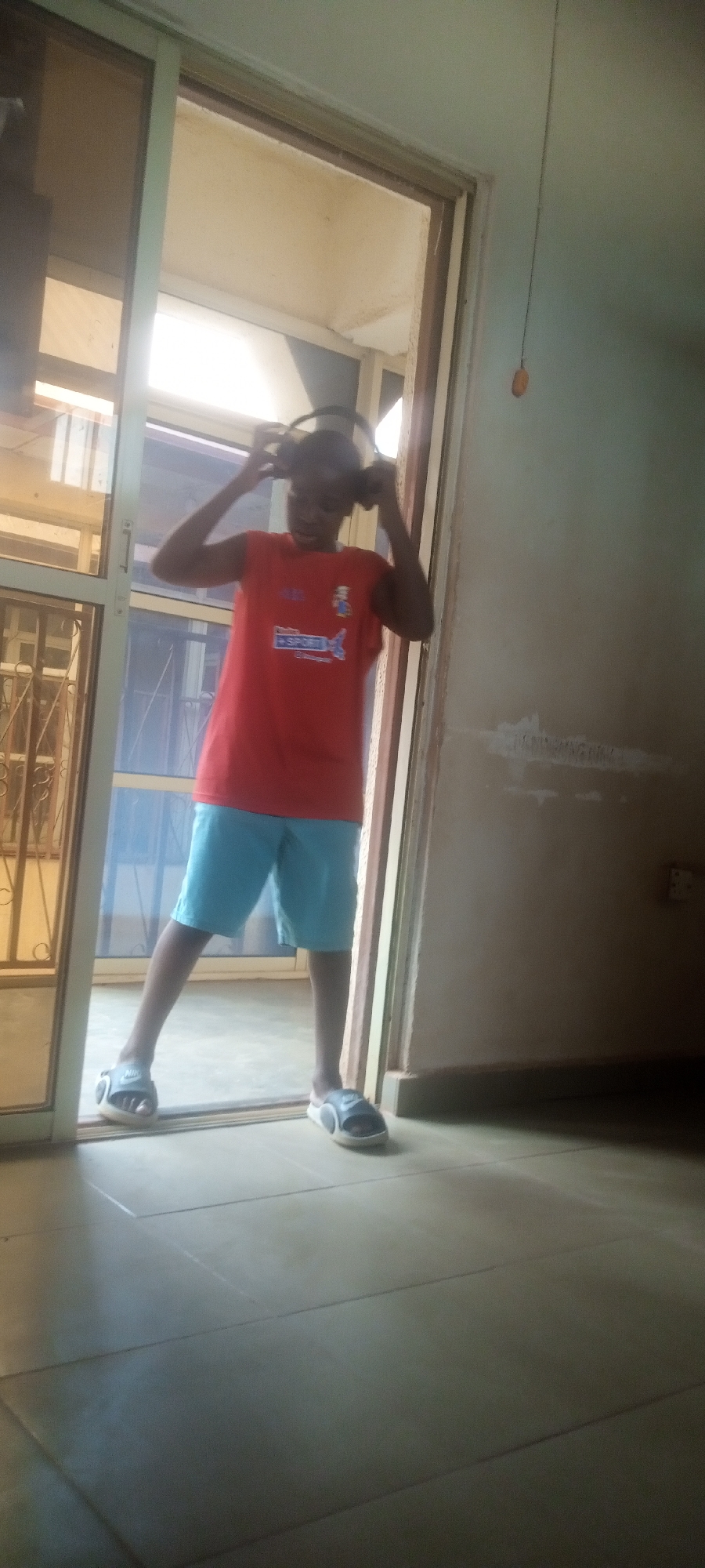 A young person, likely a teenager, is standing inside a doorway, placing headphones on their head. They are wearing a red sleeveless shirt with a blue emblem and writing on it, light blue shorts, and slip-on sandals. The doorway leads to what appears to be an enclosed porch or balcony. The interior space has tiled flooring and beige walls. An electrical outlet is visible on the wall.
The location is presumed to be Yelwa, Nigeria.