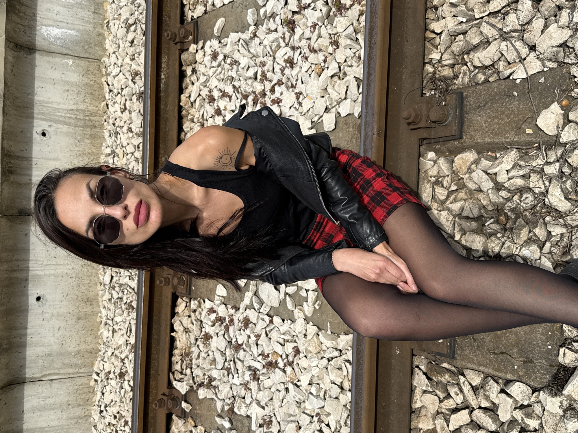 A woman with long, dark hair and sunglasses sits on a railway track bed composed of gravel and ballast, near a concrete wall in Pleven, Bulgaria. She wears a black tank top, a red and black plaid skirt, and black tights. A black leather jacket is draped over her shoulder. A small tattoo is visible on her upper arm.