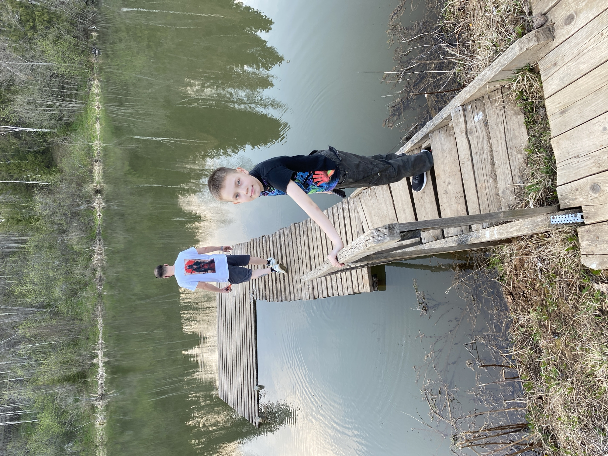Two males stand on a wooden dock extending into a body of water near Баклуши, Russia. The first male, a child, is lying prone on the dock, gripping a wooden rail. He is wearing a black t-shirt and black pants. The second male, presumed to be an adult, stands further back on the dock, wearing a white t-shirt and dark shorts. The water reflects the sky and surrounding trees. Vegetation lines the edges of the water.