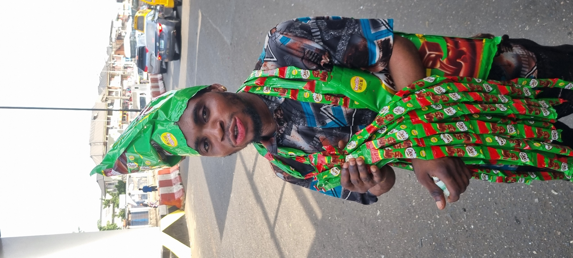 A man stands in Lagos, Nigeria, wearing a green plastic hat and a patterned shirt. He is surrounded by and holding numerous candy bars, specifically "Kellito Choco" bars. The street scene includes vehicles and buildings in the background. The day appears bright and sunny.