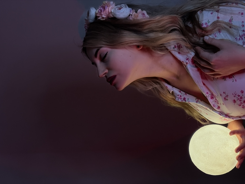 A female subject is centrally depicted in a medium close-up, facing right, with her head tilted upwards and eyes closed. Her blonde, wavy hair is adorned with a headband of light-colored artificial flowers. She wears a collared shirt with a pink floral pattern. In her outstretched left hand, she holds a glowing, spherical object resembling a moon, which emits a warm yellow light. This light, along with an additional red-pink ambient light from the left, illuminates her face and upper body, creating a gradient from yellow-orange on her lower face to pink-red on her upper face and hair. Her right hand is positioned on her upper chest. The background is a dark, indistinct plane, devoid of discernible features. This indoor scene is situated in Budva, Montenegro.