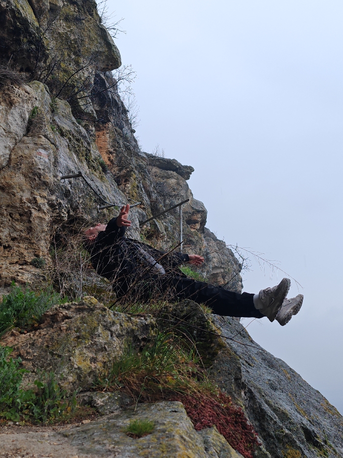 An individual is depicted lying supine on a narrow ledge or depression within a steep, rocky cliff face near Shumen, Bulgaria. The subject, wearing dark long-sleeved clothing and light-colored athletic shoes, is positioned roughly horizontally, extending outwards from the rock. Their head is oriented towards the left, with feet towards the right. The left hand is raised, displaying a two-fingered "V" gesture. Several horizontal metal bars, indicative of a secured climbing route or via ferrata, are embedded into the rock directly behind the individual's upper body. Dry, sparse vegetation partially obscures the subject's torso, while green and reddish plant growth adheres to the rock formations in the foreground and surrounding areas. The background consists of an overcast, light grey sky. The rock face itself exhibits varied textures and shades of grey and brown, with some moss or lichen.