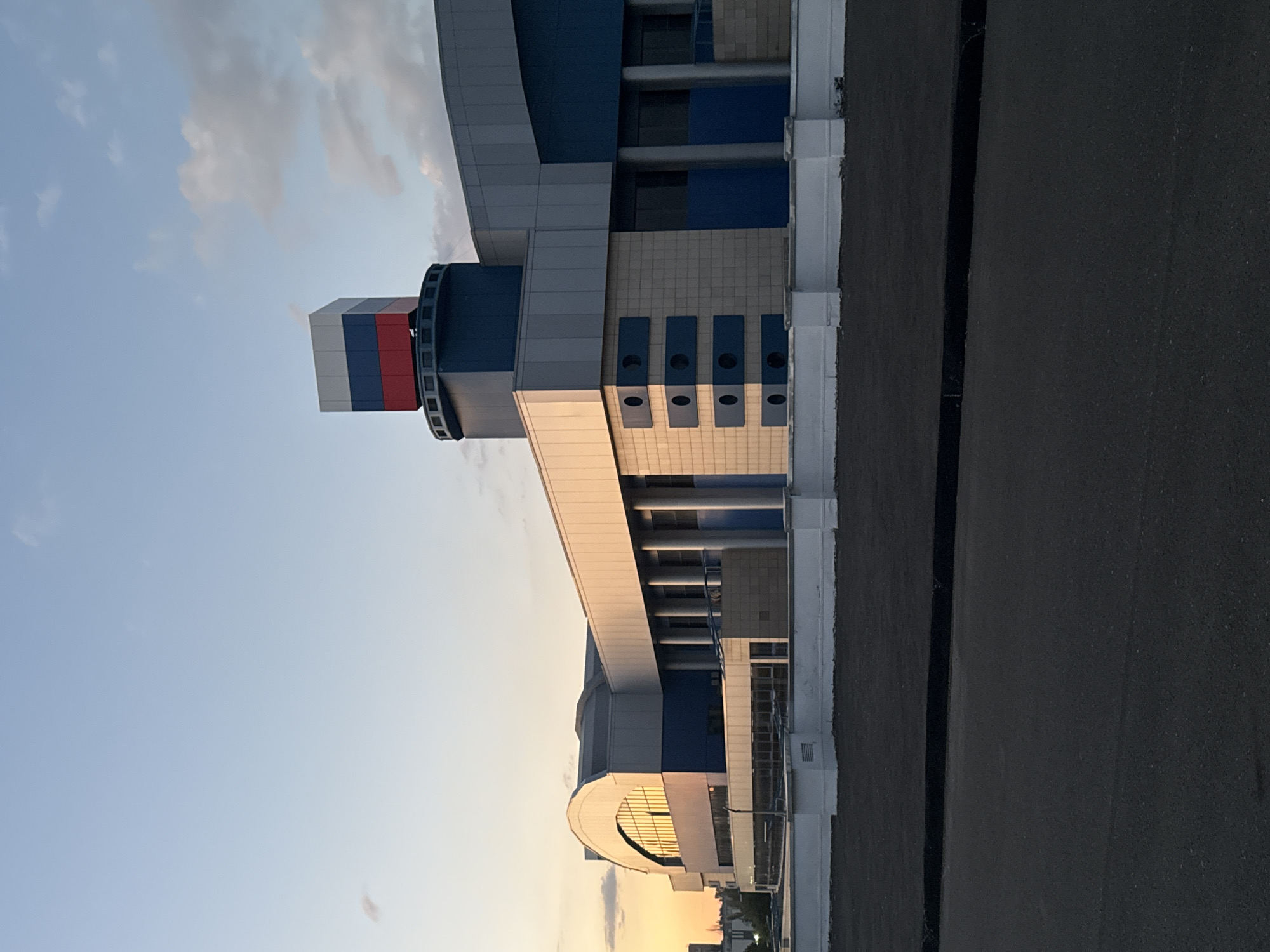 A wide, eye-level, outdoor, horizontal, full-shot photograph depicts the exterior of a modern, multi-story building in Mariupol, Ukraine, during twilight. The building's architecture is angular, with sections of white, blue, and beige cladding. A prominent feature is a cylindrical tower topped with a cube displaying the Russian flag's colors: white, blue, and red. The building has large, arched windows with reflective surfaces, and rows of circular openings are visible on one side. Tall, cylindrical columns support an upper section of the building. A low, white, walled barrier runs along the foreground. A dark asphalt road occupies the bottom of the frame, and behind the barrier is a patch of dark, possibly recently paved ground. The sky is a soft gradient of blue, transitioning to warm orange and yellow hues near the horizon, indicating sunset or sunrise, with scattered clouds. No people are visible in the scene. The overall impression is one of modern infrastructure against a dusky sky.