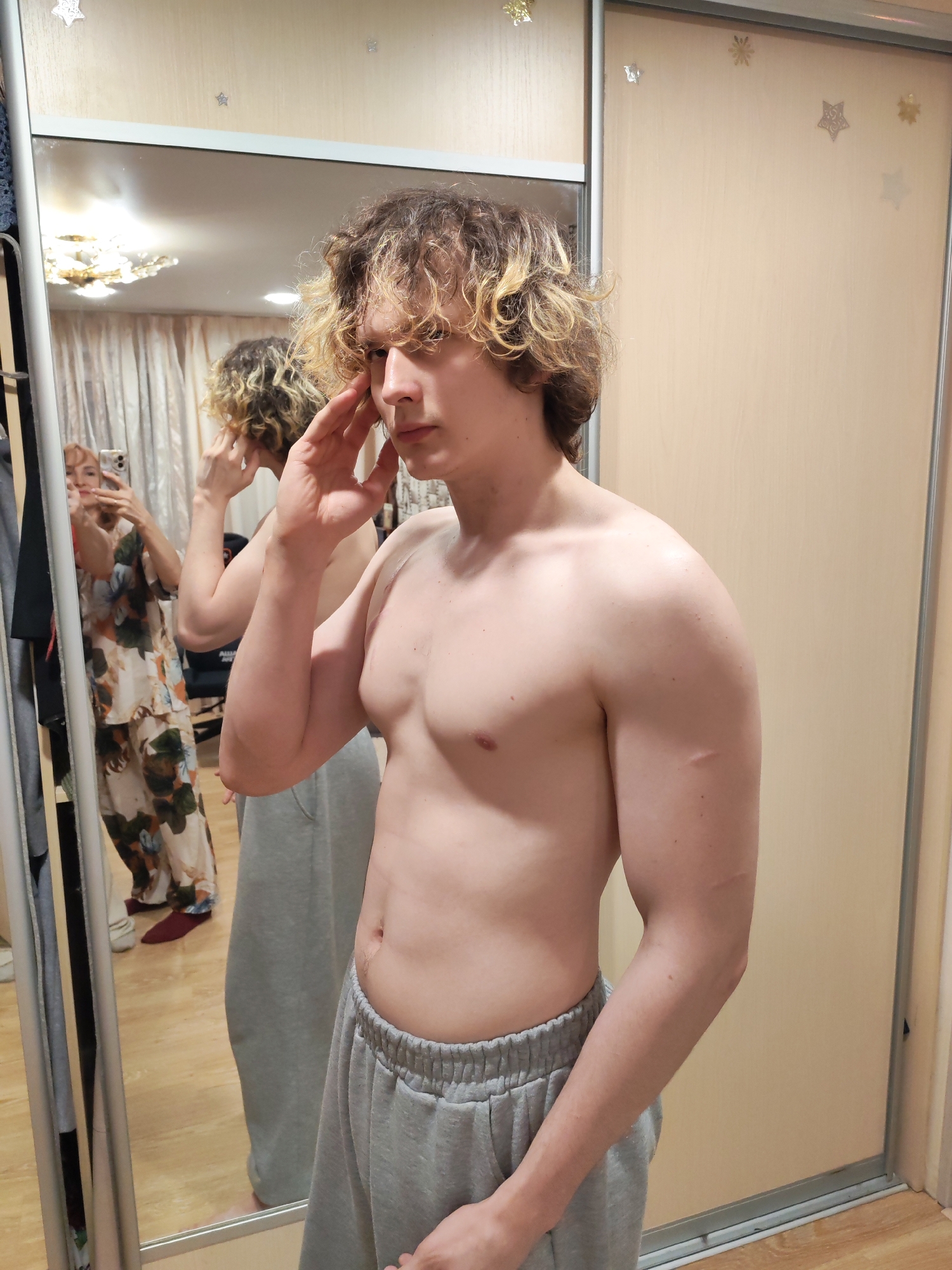 A young man with blonde curly hair, bare-chested and wearing grey sweatpants, stands in front of a full-length mirror. He is touching his face with his right hand. The mirror reflects the man and a woman in pajamas taking a picture of him with a smartphone. The scene appears to be indoors, in a room with a closet. The closet doors are a light beige color and decorated with star-shaped stickers. The setting suggests a domestic interior, possibly a bedroom or dressing area. The city of Chaikovsky, Russia is not explicitly identifiable from the image alone.