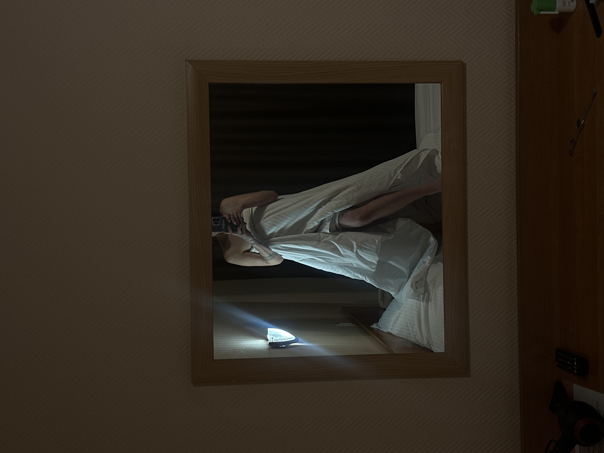 The image depicts a mirror hanging on a wall. Reflected in the mirror is a person lying in a bed covered with white linens. A phone is being held, presumably taking the picture. A small light source is visible in the reflection as well. The photograph was reportedly taken in Perm, Russia.