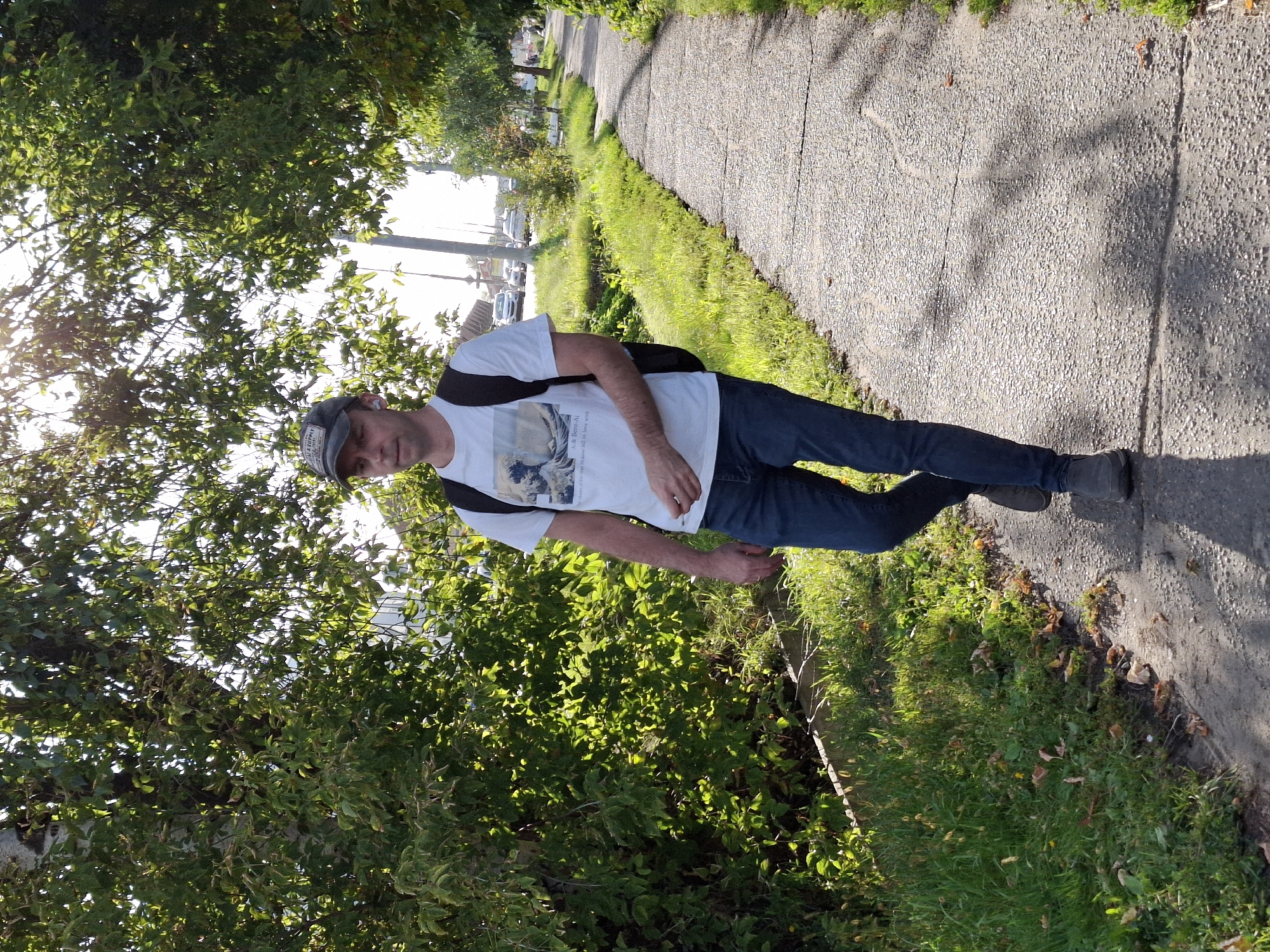 A full shot captures a man walking along a paved sidewalk next to a patch of grass and trees. The man, wearing a white t-shirt with a graphic, blue jeans, a baseball cap, and black shoes, carries a black backpack. Sunlight filters through the trees, casting shadows on the sidewalk. In the background, cars are visible on a street. The scene appears to be in a suburban or residential area.