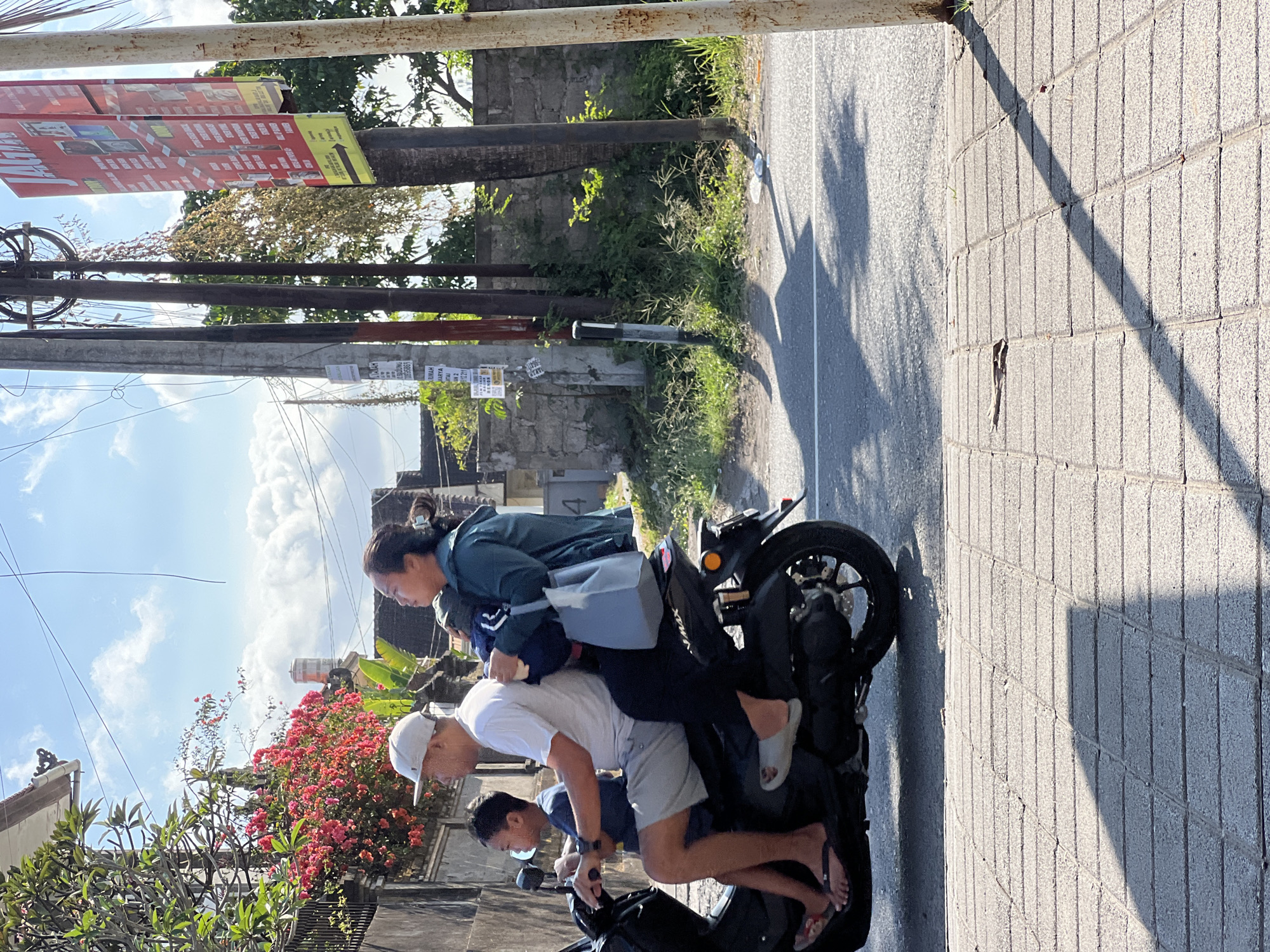 A family of three rides on a scooter on a street in Denpasar, Indonesia. The father is driving, the mother is in the middle, and a young boy is in the front. The mother is holding a baby. They are all wearing casual clothes. The scooter is black and has a license plate. The street is paved and there is a sidewalk on the right. There are buildings and trees in the background. The sky is blue with some clouds. The image appears to have been taken during the day.