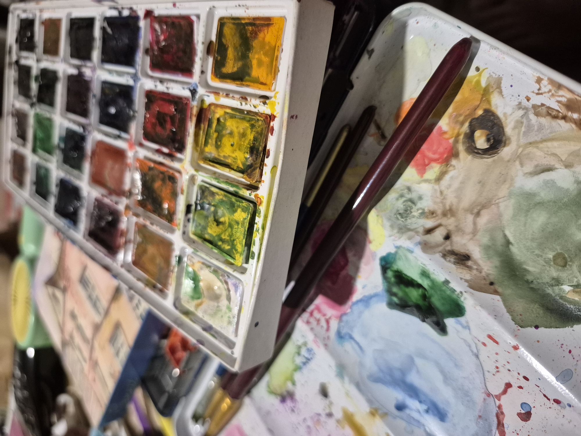 The image shows a close-up, slightly angled view of a watercolor paint set and a paint palette, suggesting an artistic or creative setting. The paint set, a white rectangular plastic case, is filled with numerous small, square pans of watercolor paint in various colors, including reds, yellows, greens, blues, and browns. Some of the paint pans appear to be wet or recently used, with visible brush strokes and water residue.
In the foreground, a white, irregularly shaped paint palette is visible, covered with splotches and blended areas of watercolor paints. Various shades of blue, green, red, yellow, and brown are present on the palette, indicating active use. Several paintbrushes, with dark red handles and metallic ferrules, are resting on the palette and extending towards the top of the image. One paintbrush is positioned prominently across the palette, partially obscuring some of the paint.
In the background, out of focus, are other art supplies. A portion of what appears to be a box or case containing more art materials is visible, with a patterned design suggesting a manufactured product. Several cylindrical objects, possibly more paint tubes or containers, are also visible. The overall impression is one of an artist's workspace, with supplies laid out for immediate use.
There are no people visible in the image. The setting appears to be indoors, possibly a studio or a dedicated art area within a home. The image does not contain any visual cues or information that would allow for its location to be determined within the city of Popovo, Bulgaria, or any other specific geographic location.