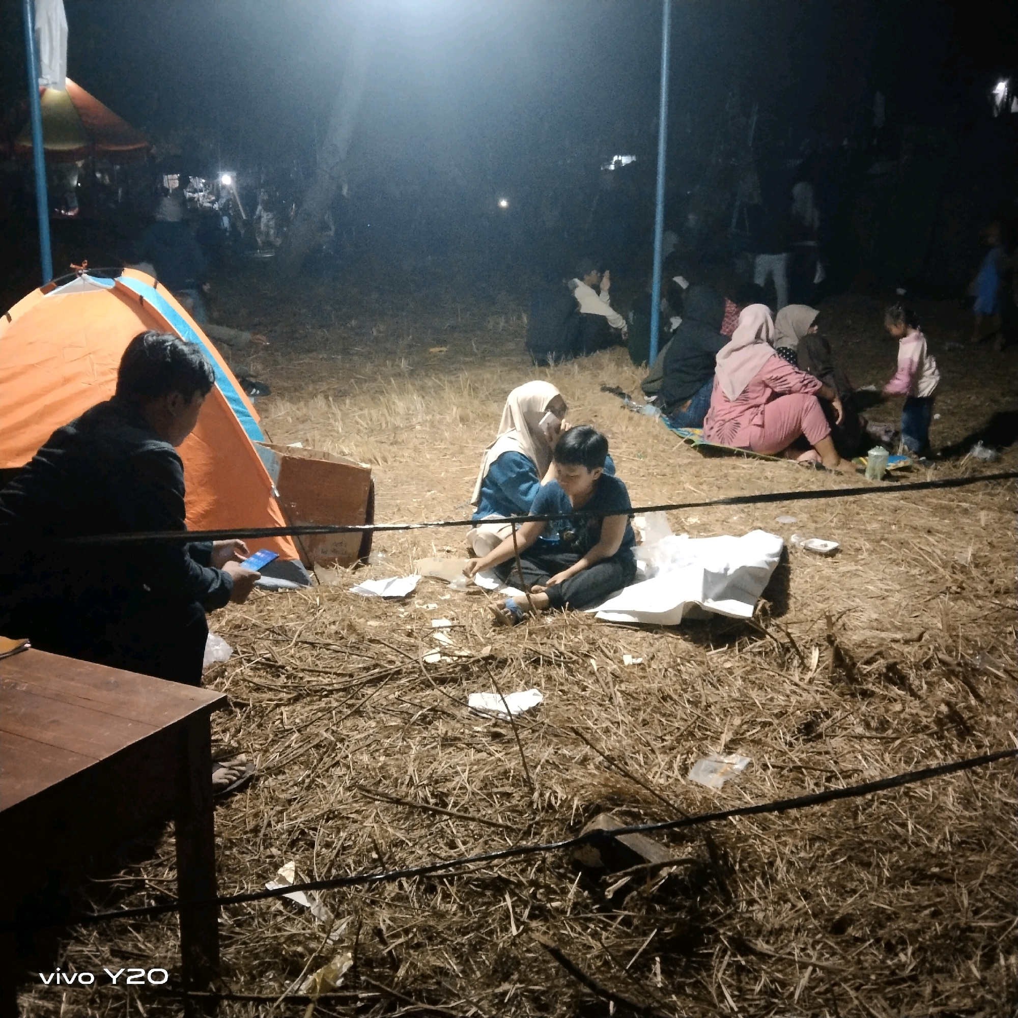 The image shows a nighttime scene at an outdoor gathering. In the foreground, a person wearing a dark jacket is seated on the ground, looking at a mobile phone. To their right, an orange tent is partially visible. In the middle ground, two children are sitting on the ground. One child is wearing a blue shirt and dark pants, and the other child, wearing a headscarf, is leaning towards the first child. Several other individuals are scattered throughout the scene, some appearing to be adults, and a young child is standing to the right. The ground is covered in dry grass and straw, and there are some scattered papers and a white object. In the background, a tent and various people are dimly visible in the low light. The overall impression is of a casual outdoor event or camping activity occurring after dark.