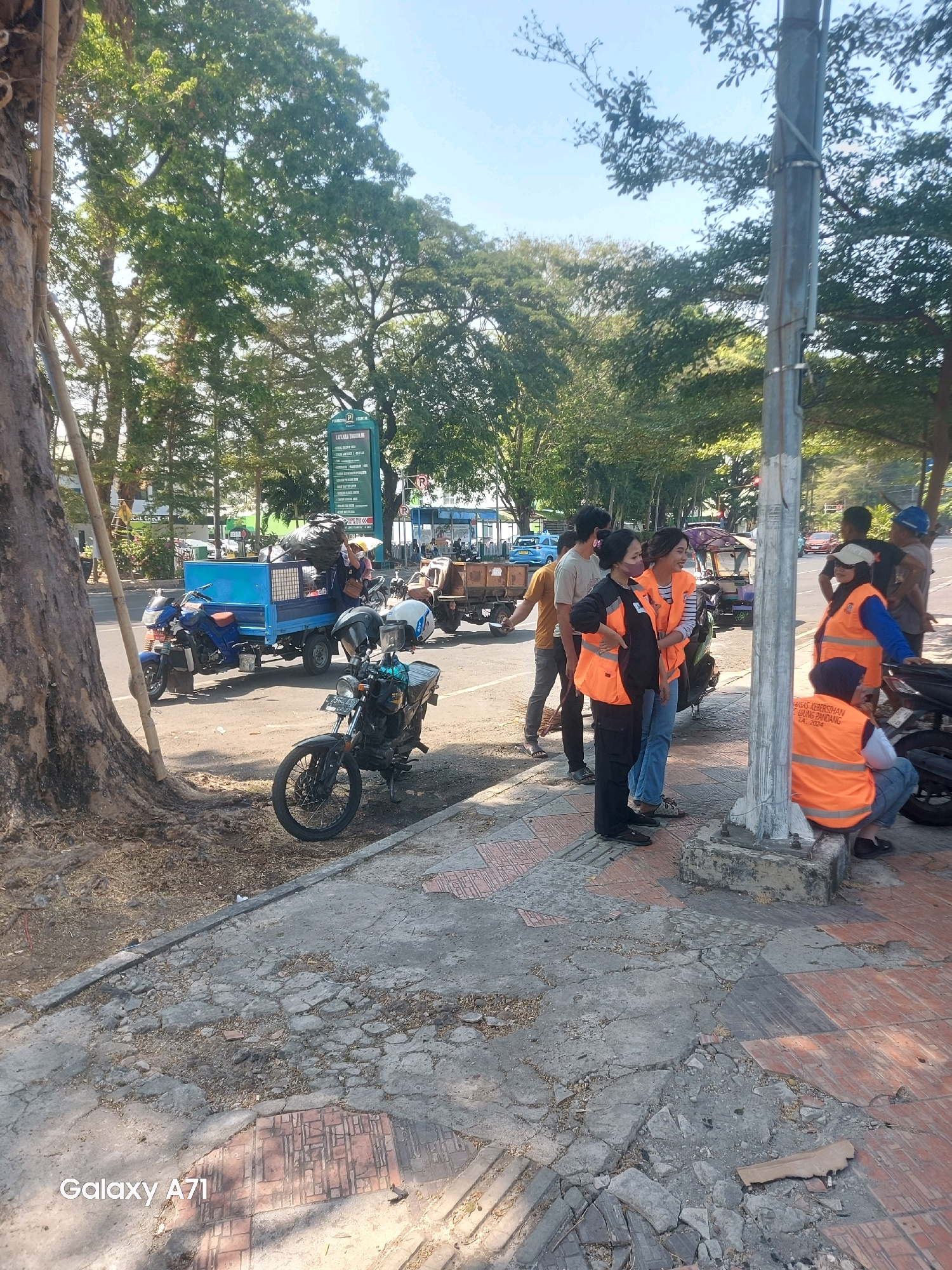 In Makassar, Indonesia, on a sunny day, a group of people, some wearing bright orange vests, are gathered on a sidewalk next to a busy road lined with trees. In the foreground, a black motorcycle is parked on the sidewalk, with a light blue three-wheeled vehicle, loaded with trash bags, driving past on the road. Further down the road, another three-wheeled vehicle with a trailer is visible. A street sign with "PELAYANAN RUMAH SAKIT" and other text in Indonesian is posted on a pole. The sidewalk is cracked and uneven.